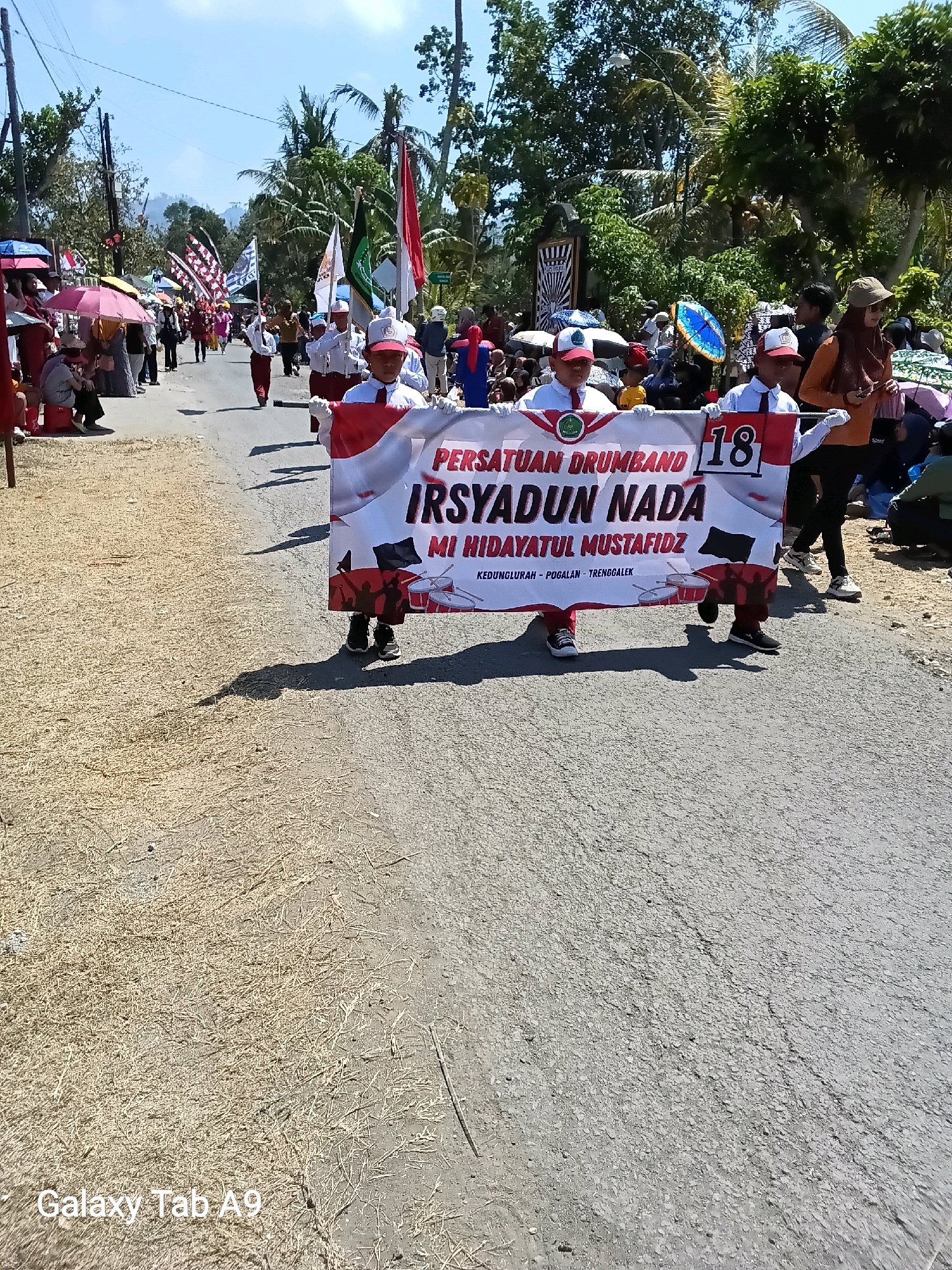 In Ngadirenggo, Indonesia, a group of children in uniform march in a parade, carrying a banner that reads "PERSATUAN DRUMBAND IRSYADUN NADA MI HIDAYATUL MUSTAFIDZ" and displays the number "18". The children are wearing white shirts with red ties and red hats, and some are holding white gloves. The banner has an image of drums and silhouettes of marching figures. The parade is taking place on a paved road under a clear blue sky. Spectators line the sides of the road, some holding umbrellas for shade. The environment includes trees, buildings, and a mountain in the background. The ground on the left side of the road is covered with dry straw or hay. The "Galaxy Tab A9" watermark is visible in the lower left corner of the image.