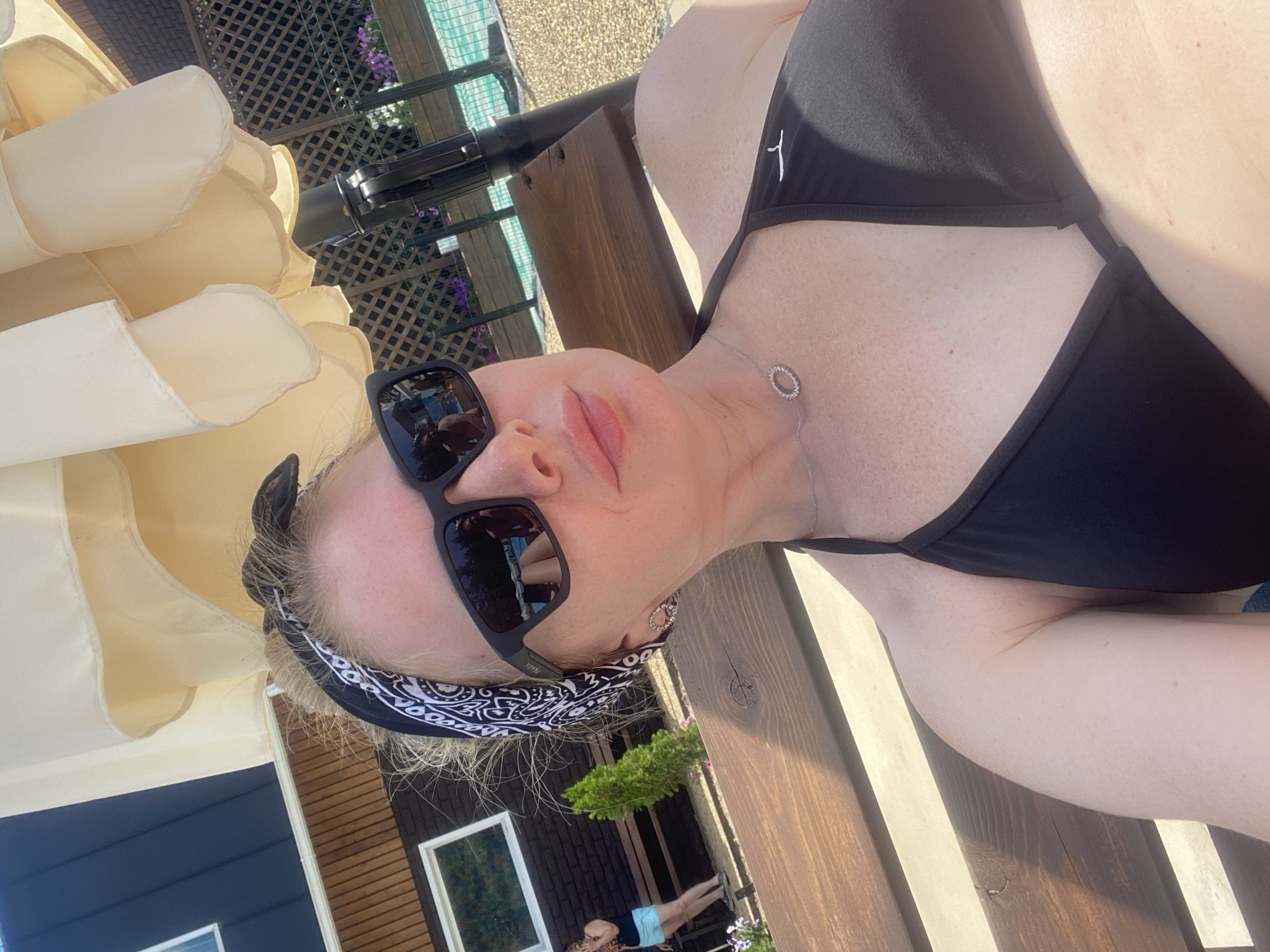 A Caucasian woman with blonde hair tied back with a black and white bandana, wearing a black bikini top, black sunglasses, and a silver necklace, is seen outdoors in a close-up, low-angle shot. She is seated on a wooden bench under a large, cream-colored umbrella. In the reflection of her sunglasses, a man can be seen sitting on a lounge chair near a swimming pool. The background shows a building with dark siding and a white window, a wooden fence, and a swimming pool area with landscaping. The overall setting appears to be a recreational area, possibly a resort or a private backyard, during daylight hours. The location is identified as Voronezh, Russia, but this information is not visually verifiable from the image itself.