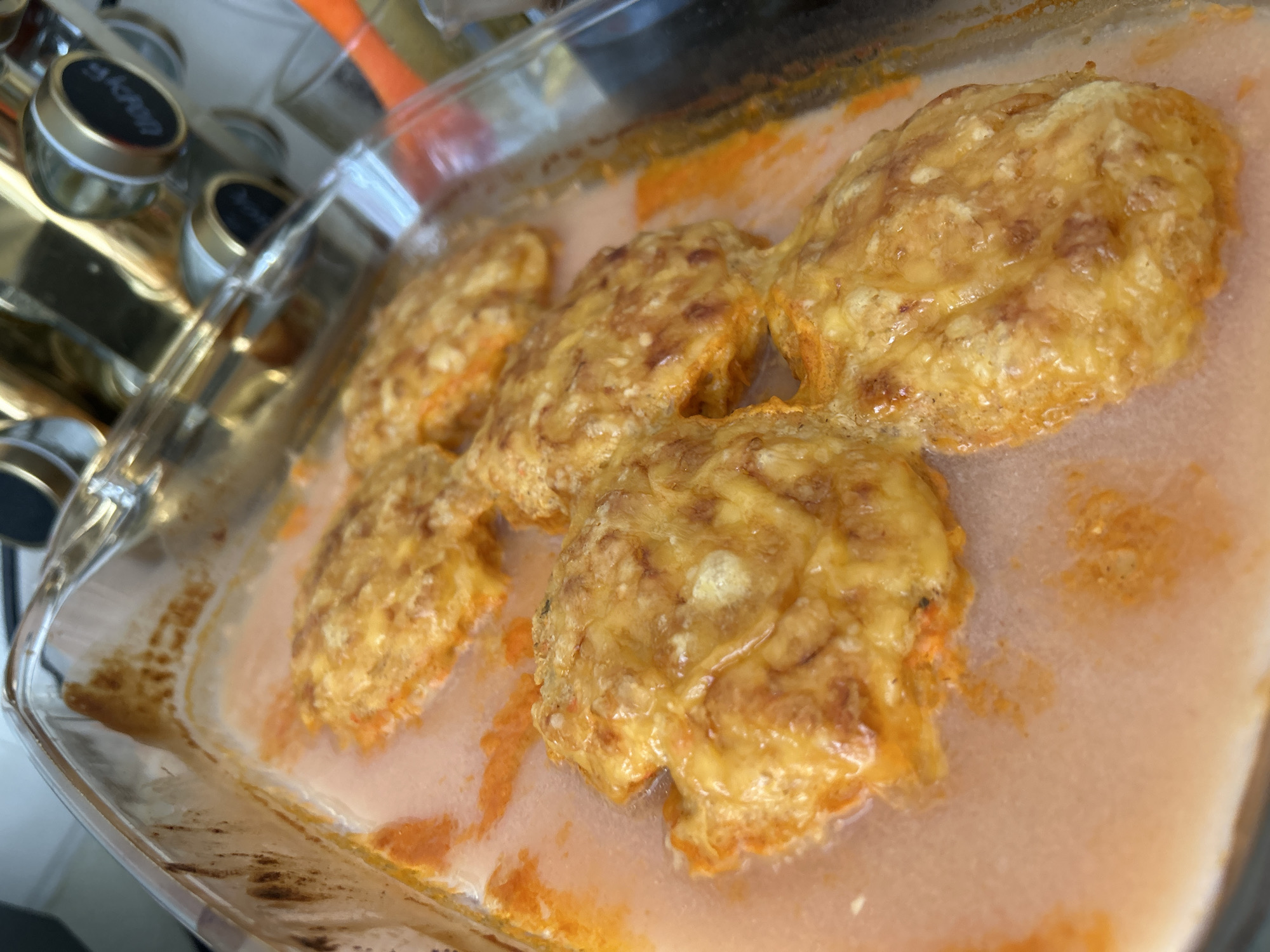 In a clear glass baking dish, five golden-brown, cheese-topped food items are submerged in a light orange broth. The food items, appearing to be some form of patties or cutlets, have a slightly irregular, rounded shape with visible texture from the cheese and possibly breadcrumbs. The broth is a pale pinkish-orange and a few small specks of orange solids are visible dispersed within it. The baking dish itself has some light brown residue along the upper edges, suggesting it has been used for baking. In the background, out of focus, are several spice jars with metallic lids, some of which have black labels. An orange-handled kitchen utensil is also partially visible to the right of the baking dish. The overall environment appears to be a kitchen setting. The image is taken from a slightly elevated, angled perspective, looking down into the dish. There are no people visible in the image, nor any indication of the specific location within Noviy, Russia.
