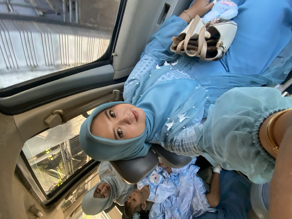 The scene depicts the interior of a vehicle in Tambakrejo, Indonesia, occupied by three individuals. In the foreground, an adult female with fair skin is positioned, looking directly at the camera. She wears a light blue hijab and a matching long-sleeved dress with white, bird-shaped appliques and white beads on the bodice and sleeves. A gold-colored bracelet is visible on her left wrist. A beige handbag with a brown strap and a light blue fluffy object (possibly a keyring with a rabbit character) rest on her lap. Behind her, another adult female is visible in a light grey hijab and top, looking towards the front. Further back and to the left, a young child with a blue hair clip is seated, wearing a light blue and white outfit featuring a character resembling Elsa. The vehicle's interior features light-colored trim and upholstery. Through the windows, blurred elements of an outdoor environment, including vegetation and a vertical slatted structure, are visible, suggesting an urban or suburban context.