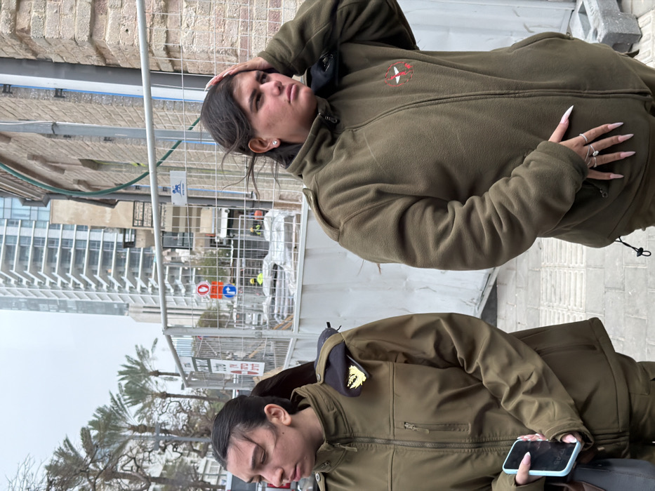 This image depicts two young adult females in olive-drab military-style jackets standing on a paved sidewalk in an urban setting in Tel Aviv, Israel, under an overcast sky.
The female on the left wears a zippered, olive-drab softshell jacket with a black beret and a gold insignia tucked under her left shoulder epaulet. Her dark hair is pulled back, and she holds a light blue smartphone in her right hand, her expression neutral as she gazes slightly to her left.
The female on the right wears a zippered, olive-drab fleece jacket with a red and white circular patch (resembling an emblem) on her right chest. Her dark hair is loose, appearing wind-blown, and she has her left hand resting on her forehead. Multiple rings are visible on her left hand's fingers. Her expression appears contemplative or slightly pensive. Both individuals have long, light-colored fingernails.
Behind the individuals, a grey metal construction barrier separates the sidewalk from an active construction site. Visible within the construction zone are excavated earth, large pipes, various building materials, and at least two workers wearing yellow vests and hard hats. Traffic signs, including a red circular 'no entry' sign and a blue circular 'turn left' sign, are also present. An older, light-brown brick building with a green pipe running vertically on its exterior is situated to the right of the construction site. In the far background, a tall, modern, multi-story glass and concrete skyscraper is prominent, alongside palm trees and other deciduous trees lining a street.