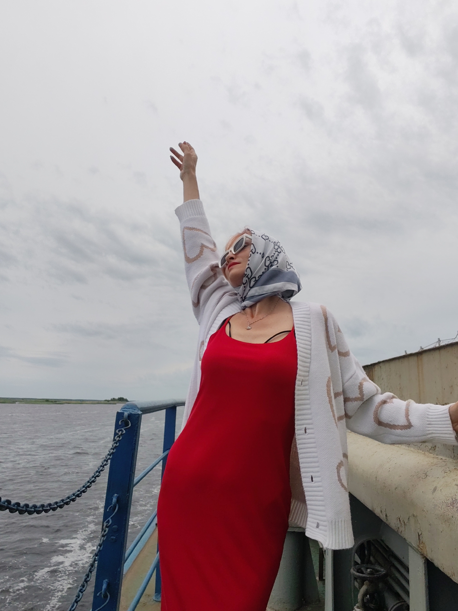 A woman stands on the deck of a boat, her right arm raised towards the sky, her fingers spread. She is wearing a red dress and a white cardigan with a heart pattern, a patterned scarf on her head, and white sunglasses. The scene appears to be a cloudy day, with a body of water on the left and a railing on the right. The woman's pose suggests a sense of freedom or exhilaration. The location is identified as Bereg, Russia.