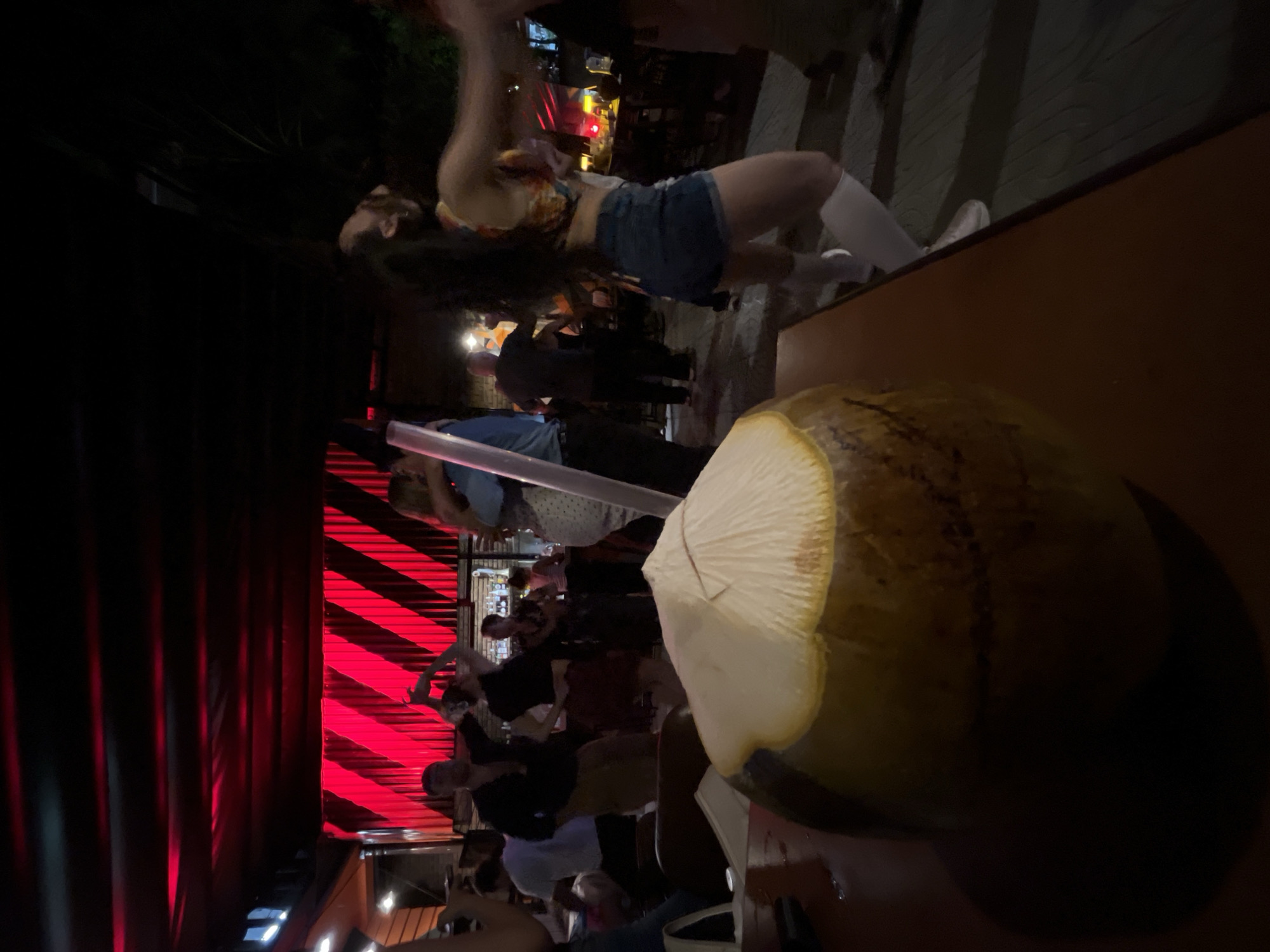 The scene, described as being in Đà Nẵng, Vietnam, depicts a lively nighttime bar or club environment. In the immediate foreground, a full coconut beverage, with its top cut open and a white straw inserted, rests on a reddish-brown wooden table, casting a dark, crescent-shaped shadow.
Beyond the table, numerous individuals are present, engaged in social activities and dancing. On the right, a woman with long dark hair, wearing a patterned crop top, blue denim shorts, and white knee-high socks over white shoes, is captured mid-movement with her arms raised, suggesting dancing. In the center-left, a man in a dark shirt and light brown pants is dancing with a woman in a dark top and reddish skirt. Other patrons are visible throughout the midground and background, standing, mingling, and dancing.
The environment is characterized by distinctive red lighting. The ceiling features dark panels with linear red strip lights. A prominent background wall is illuminated with intense red light, highlighting its corrugated or slatted texture and creating a pattern of diagonal red stripes. In the far background, a bar counter with bottles and additional lighting is discernible. The floor surface appears dark with a visible linear pattern. The overall atmosphere is dark and vibrant, with the red lighting being a dominant visual element.