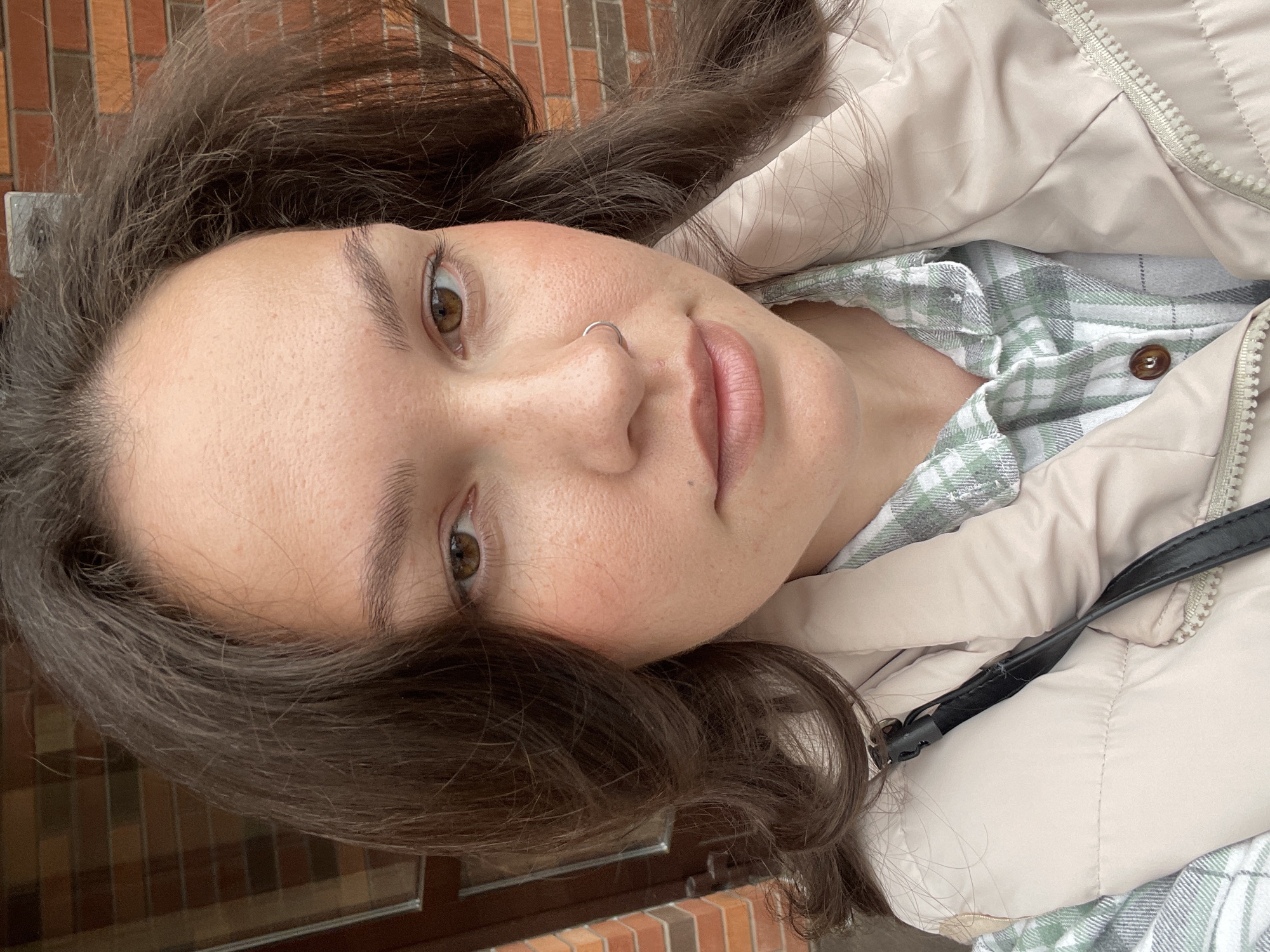 A close-up, tilted, slightly downward-angled shot features a young woman's face. She has brown hair, brown eyes, fair skin with visible pores and freckles, and a nose ring. She wears a light-colored padded jacket over a plaid shirt. The background reveals a brick wall, suggesting an outdoor setting in the city of Izhevsk, Russia. A black strap is visible near her shoulder.