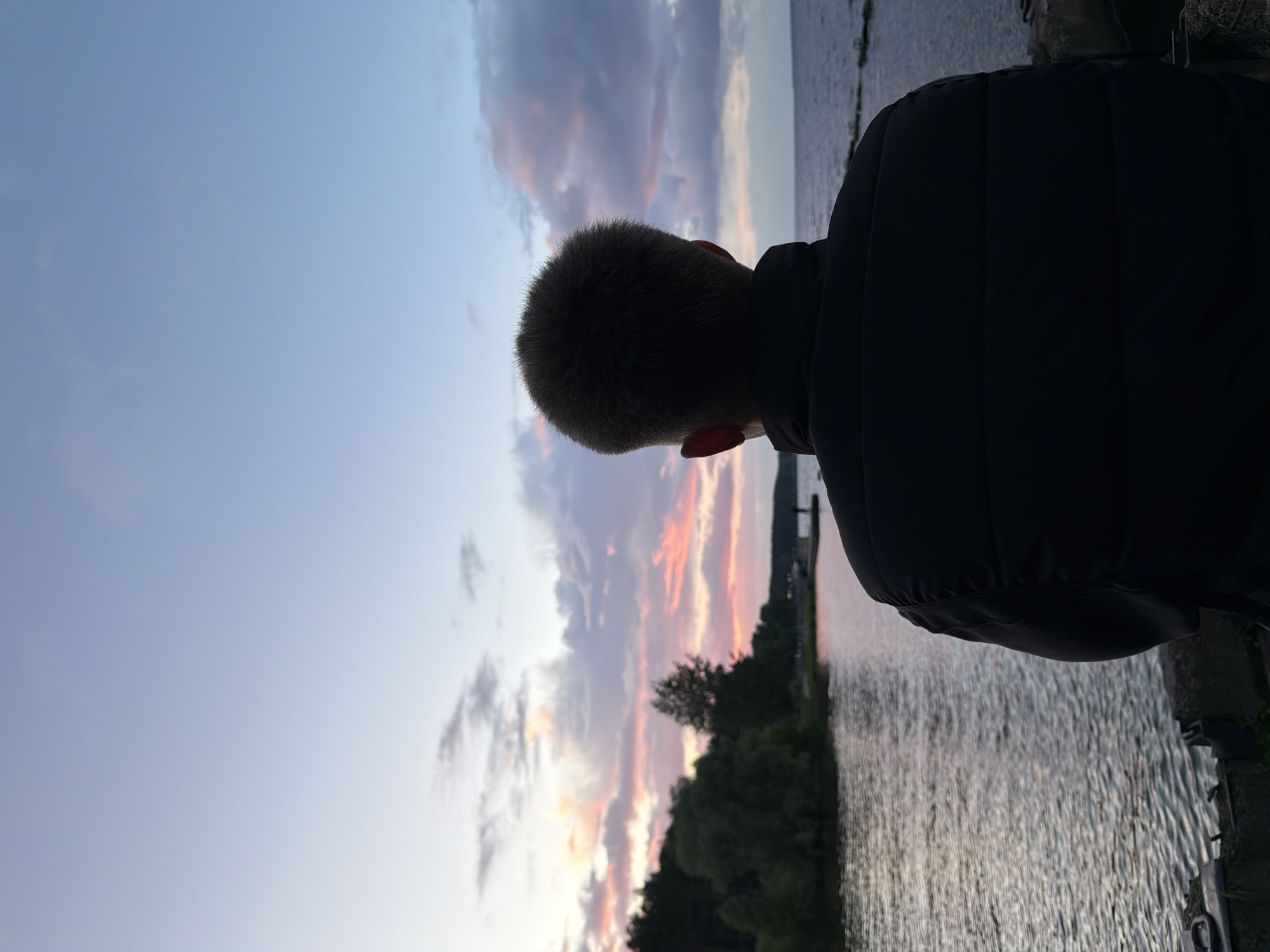 A full-shot, eye-level, vertical photograph shows the back of a young person wearing a dark puffer jacket, looking out at a body of water during sunset. The person's head is in the upper right quadrant of the image. The water reflects the colorful sky, which is a mix of soft blues, pinks, and oranges from the setting sun. Clouds are visible in the sky, some catching the light of the sunset. On the left side of the image, a tree line with dark green foliage borders the water. The right side of the image shows the open water. The foreground consists of a concrete embankment or wall. The overall scene evokes a sense of tranquility and contemplation. The location is identified as Chaikovsky, Russia.