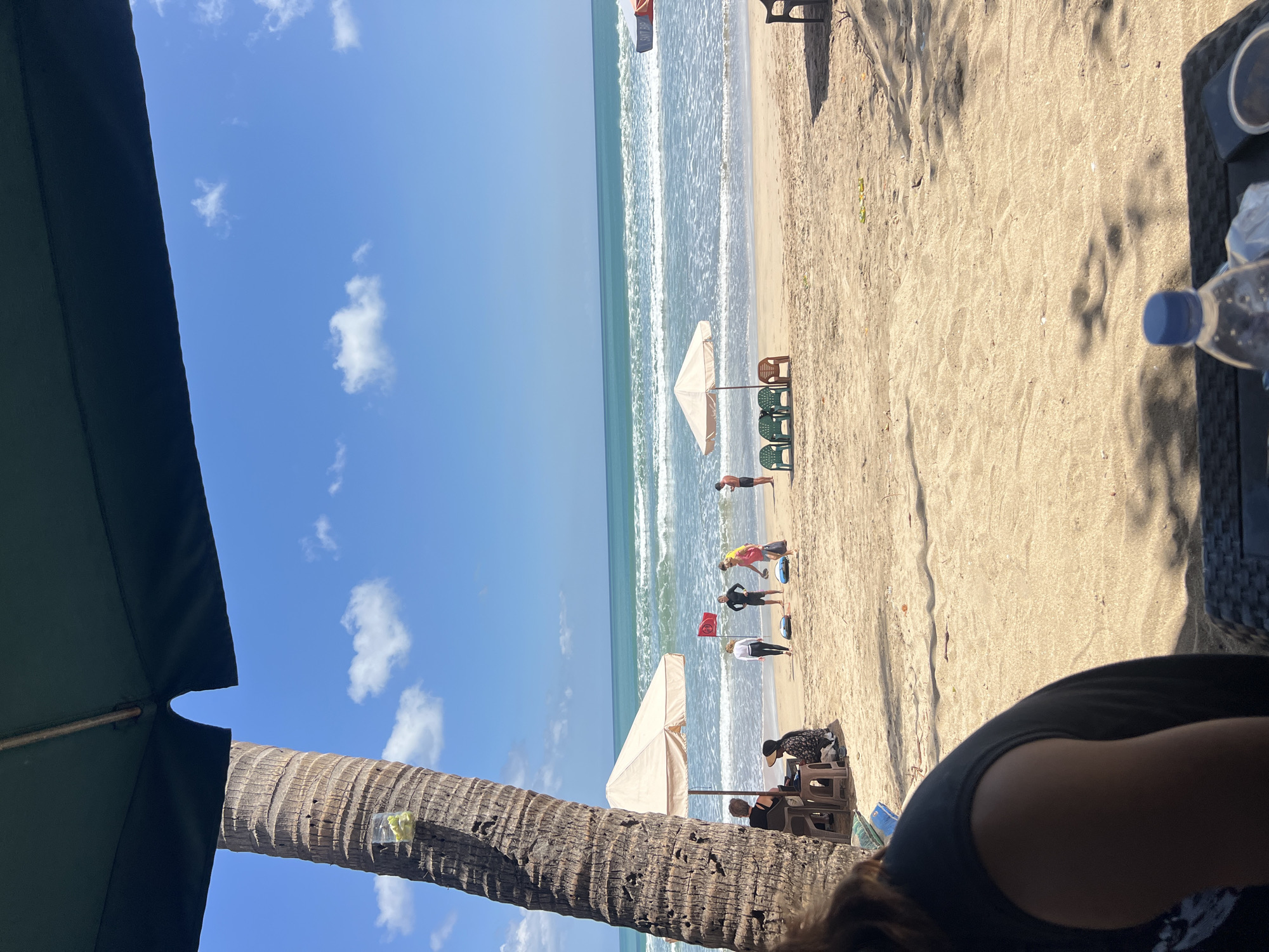 In Kuta, Indonesia, a beach scene unfolds under a bright, sunny sky with scattered white clouds. The foreground features the partial view of a person from behind, seated at a table partially obscured by shadows. A plastic water bottle with a blue cap sits on the table. A large palm tree trunk, textured and rough, dominates the left side of the frame, with a clear plastic cup containing some yellow objects tied to it.
Further back, a sandy beach stretches towards the turquoise ocean, where gentle waves create white surf. Several people are visible on the beach. To the left, under a light-colored umbrella, two women are seated. Nearer to the water, a group of four individuals are engaged in an activity, possibly a surf lesson, with one person holding a red flag. Two white beach umbrellas are set up, and a row of green and brown plastic chairs are placed on the sand. In the distance, another umbrella, orange and white, is visible, along with a silhouette of a person standing. The overall atmosphere is that of a relaxed day at the beach.