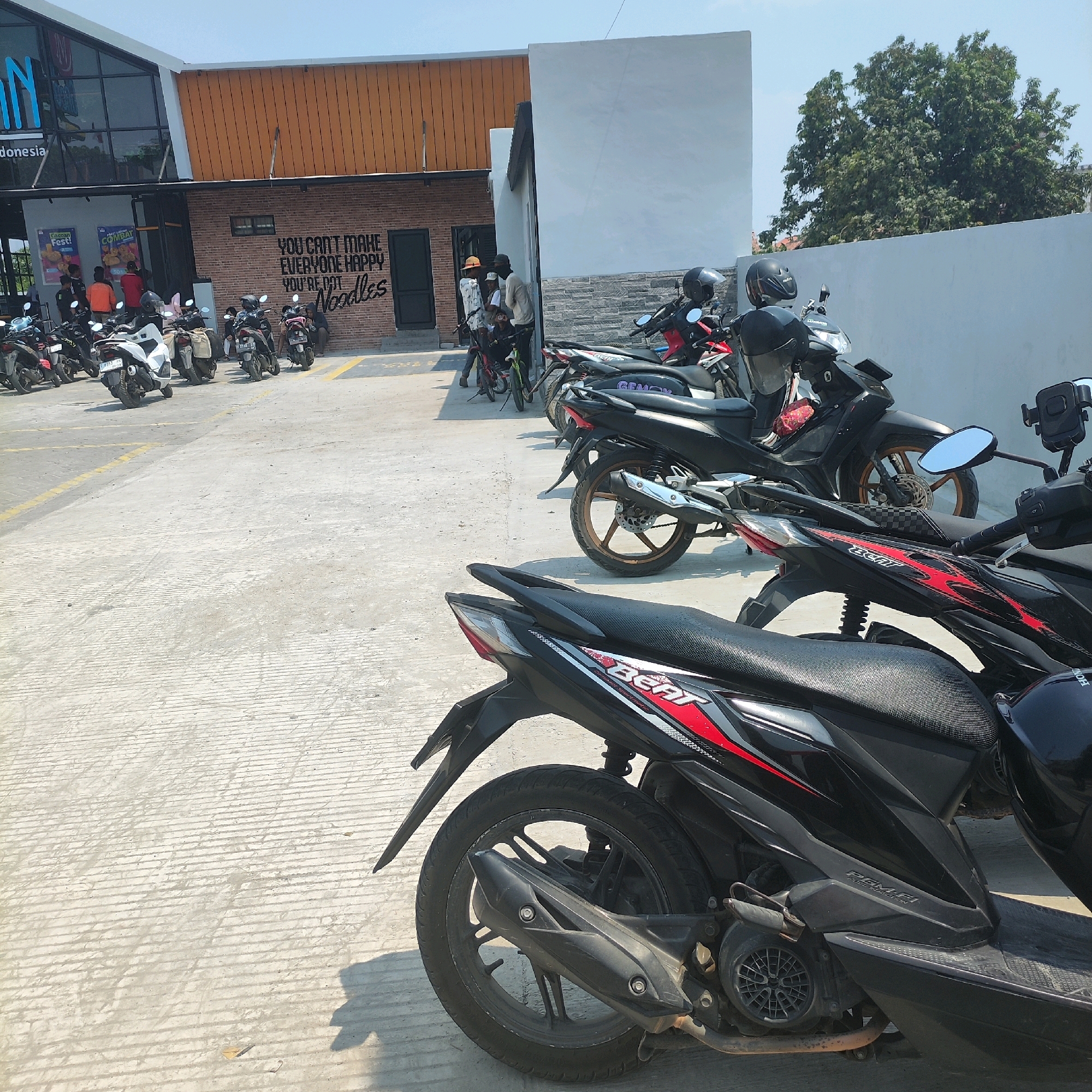 The image shows a street-level view of a parking lot area next to a modern building with orange and brick accents. The ground is a light-colored concrete with visible lines from its construction. A row of parked motorcycles, mostly scooters, occupies the foreground and middle ground, with their fronts facing away from the viewer. One black scooter with red and white racing stripes, labeled "BEAT," is prominently featured in the lower right. The building has large glass windows and a prominent sign that appears to be illuminated. There are a few people visible near the building entrance, some standing and some possibly interacting with the parked motorcycles. The sky is clear and blue, suggesting a sunny day. In the background, a lush green tree is visible, contrasting with the urban setting.