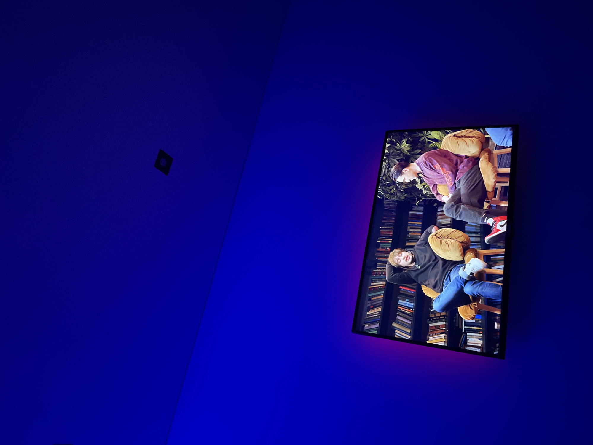 The image displays a modern, dimly lit interior space in Kazan, Russia, characterized by walls illuminated in a deep, vibrant blue hue. The walls converge at an angle, creating a distinct corner. In the upper left portion of the blue room, a small, dark, square object with a circular center is affixed to the wall.
A large, rectangular flat-screen display is mounted on the right wall, tilted diagonally. This screen is a focal point, radiating a subtle purple-pink glow onto the adjacent blue surface. The display itself presents an indoor scene featuring two young men amidst a backdrop of extensive bookshelves packed with numerous volumes, and some green foliage at the top.
The upper man on the screen has dark hair and wears a purple and maroon patterned long-sleeved shirt, dark trousers, and red sneakers. He is captured in motion, appearing to lean over or rise from a wooden chair adorned with light brown, plush cushions. The lower man, with lighter hair, is dressed in a dark hoodie, blue jeans, and light-colored sneakers. He is seen in a relaxed pose, reclining on a similar wooden chair with light brown cushions, with his hands clasped behind his head. The scene on the display suggests a casual or relaxed interaction between the two individuals. The striking contrast between the intensely blue-lit environment and the warmer, detailed scene on the screen gives the impression of an artistic installation or a contemporary setting.