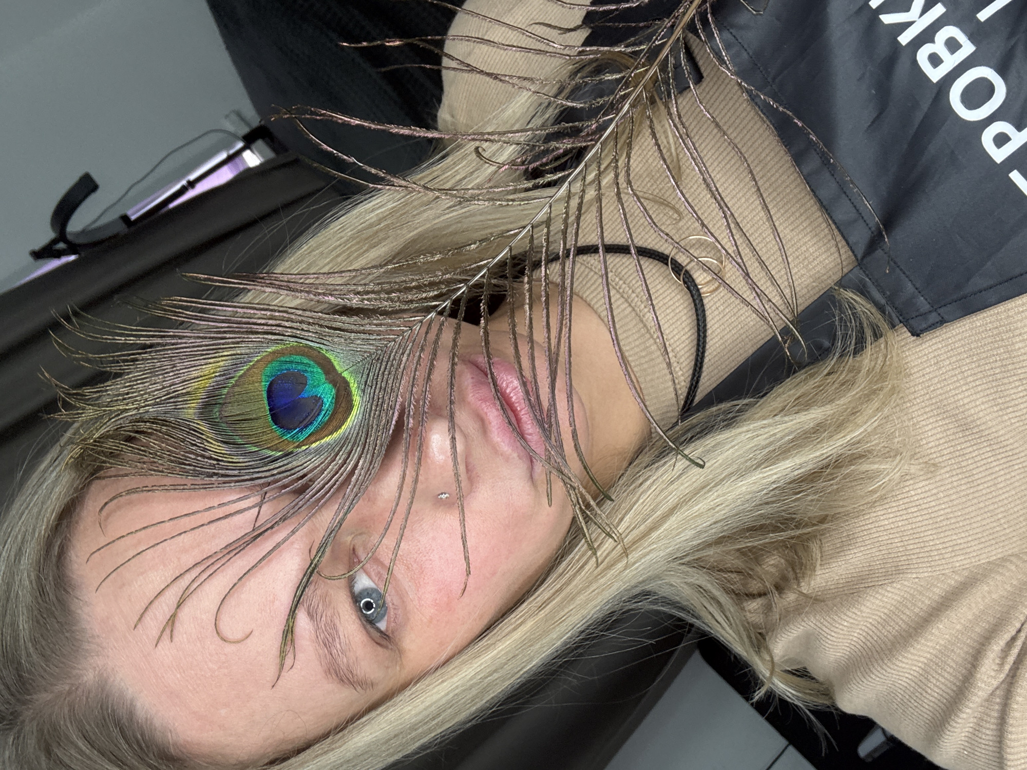 An artistic portrait captures a person, likely a woman, with long blonde hair, partially obscured by a large peacock feather. The person is seen from an oblique, almost overhead, angle, appearing to lie down or have their head tilted significantly to the left. One vivid blue-grey eye is visible, with a small facial piercing (a philtrum or medusa piercing) directly below the nose. The lips are slightly parted.
A prominent peacock feather extends across the person's face, its iridescent blue and green "eye" pattern covering one eye, while the delicate fronds of the feather fan out across the forehead and cheek. The person is wearing a light brown, ribbed long-sleeved top. To the right, a black garment, possibly an apron or bag, is visible, on which white capital letters spell "POBK". A black cord necklace with two small gold rings rests on the person's neck. In the blurry background, a pair of glasses is faintly visible, reflecting some light, suggesting an indoor setting with artificial illumination, possibly a studio. The overall mood is composed and artistic, emphasizing the interplay between the human subject and the natural beauty of the feather.