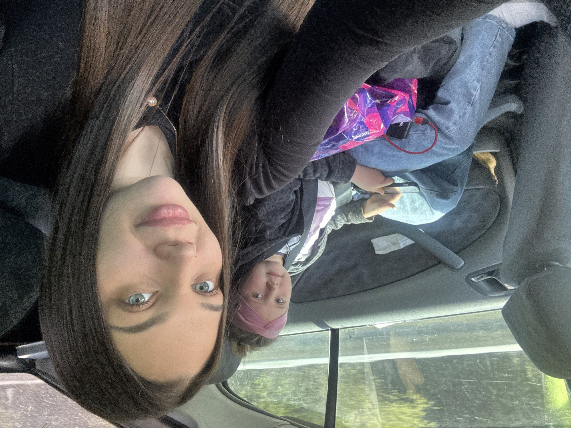 Two people, a woman and a child, are in a car. The woman has long brown hair, fair skin, and light-colored eyes, and is wearing a dark-colored top. The child is wearing a pink headband, a jacket, and jeans. They are in the back seat of the car, with a purple and pink Taco Bell bag on the child's lap. A cell phone with a red charging cord is on top of the bag. Trees are visible through the car window, indicating they are driving. The location is not specified as within the city of Krasnoyarsk, Russia.