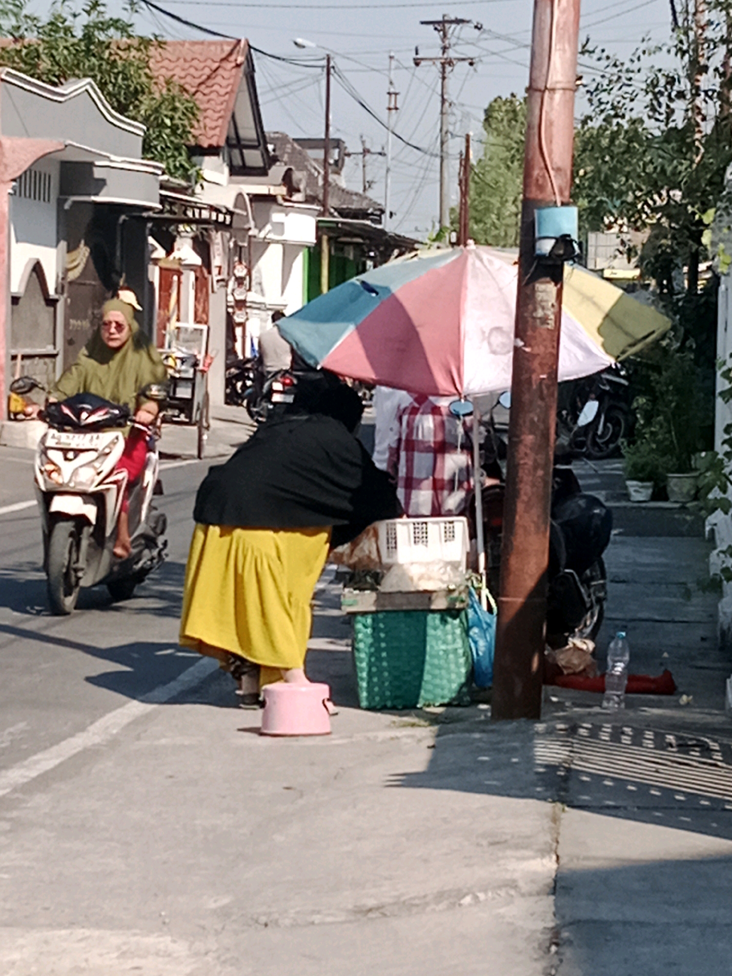 In Cemani, Indonesia, a woman wearing a green hijab and a green long-sleeved shirt rides a white scooter on a paved road. She is looking towards the left. To the right of the road, a street vendor, wearing a black headscarf and a yellow skirt, stands behind a display of goods under a colorful umbrella. The vendor is bent over, attending to her merchandise. A pink plastic stool is on the ground in front of the vendor. The environment appears to be a residential or commercial street with buildings and utility poles visible in the background. The overall scene depicts a typical day in a bustling Indonesian town.