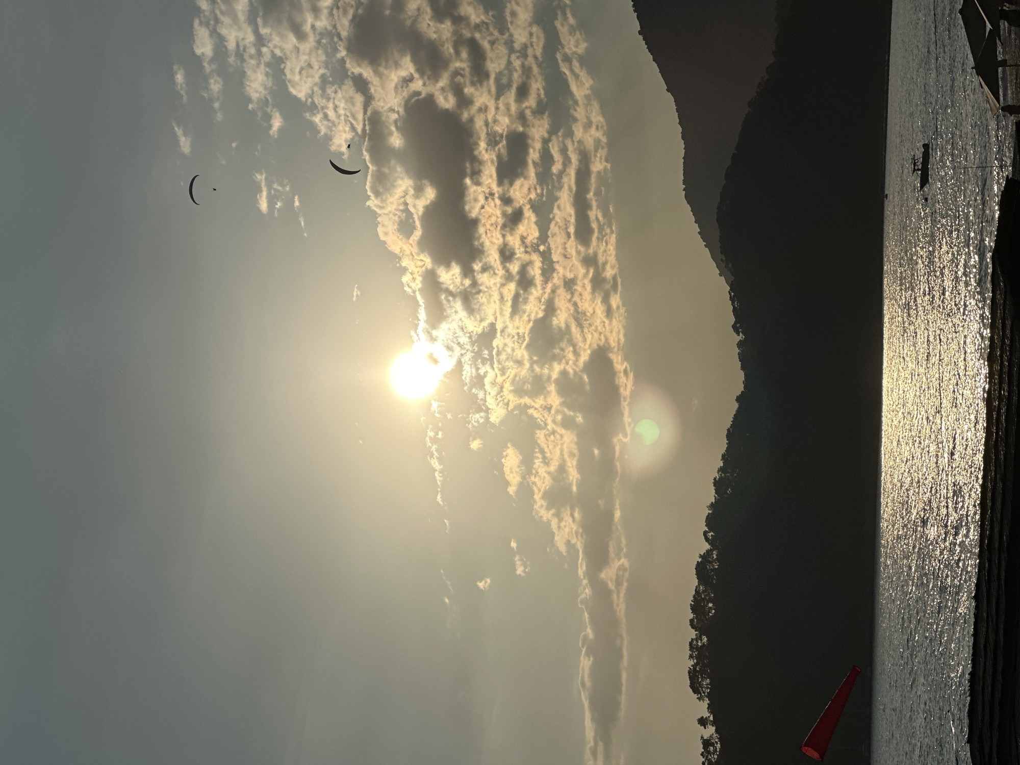 The image shows a vertical landscape of a scene in Fethiye, Turkey. The sun is high in the sky, partially obscured by clouds. Two paragliders are visible in the sky. A mountain range is on the right side of the image. Below the mountain, water reflects the sunlight. A red windsock is at the base of the mountain. A partially visible dock or structure is on the far right.