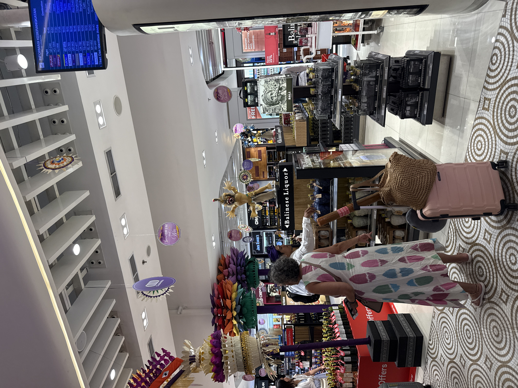 Inside a brightly lit airport duty-free store, two women are seen browsing among shelves stocked with liquor. The scene is bustling with products and various decorative elements. One woman, with curly grey hair, wears a colorful patterned dress in white, pink, blue, and green hues, and is pointing towards a display while standing next to a light pink rolling suitcase and a woven straw bag. Another woman, wearing a white shirt and a blue headscarf, stands behind a counter, appearing to be an employee or assisting the shopper.
The store features numerous displays of "Balinese Liquor" and other brands like "JACK DANIEL'S," "CHIVAS REGAL," and "MY MARTIN." Many products are labeled with "BALI." Large, colorful artificial flower arrangements and traditional Balinese decorations, including a statue resembling a mythical figure, adorn the shopping area. The floor is tiled with a distinctive circular pattern in brown and white.
A prominent digital departure information screen in the background displays "INFORMASI KEBERANGKATAN" (Departure Information) with flight details, times ranging from 16:45 to 19:40, and destinations including Phuket, Singapore, Mumbai, Perth, Doha, Abu Dhabi, Hongkong, Bangkok, Delhi, Kuala Lumpur, and Guangzhou. The text "Tiket Dan Identitas Anda" (Your Ticket and Identity) is also visible. Other visible text includes "Bali Festival," "Buy 1 get 1 FREE," "Sweeten your journey with exclusives," "Save 15%," "Spend IDR 1 M and Win," and "Special Offers." The numerous references to Bali and the Indonesian language on the departure board suggest the location is likely a duty-free shop at an airport in Bali, Indonesia, during the late afternoon or early evening.