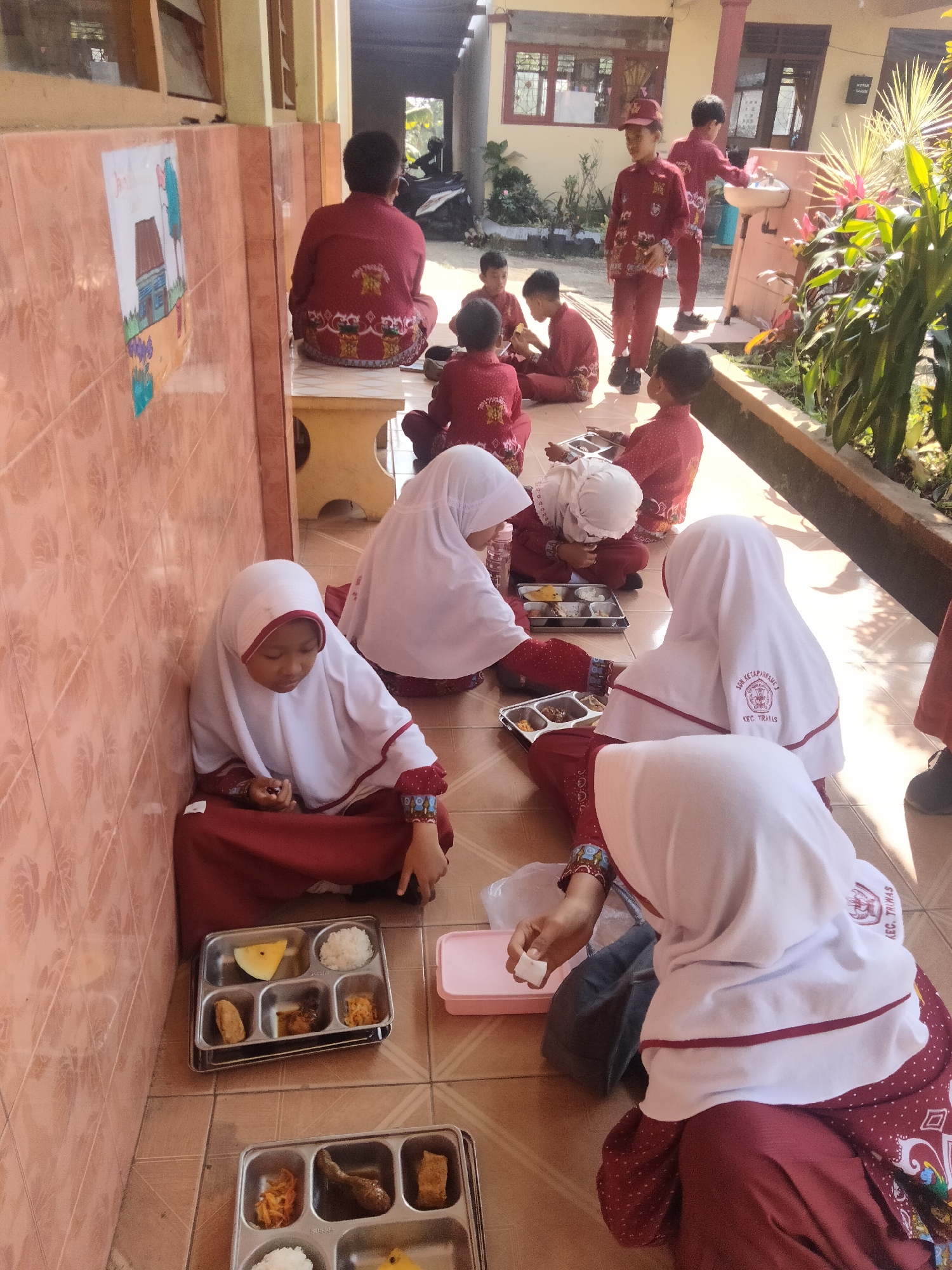 In Tretes, Indonesia, several children in maroon uniforms and white hijabs are seated on a tiled floor, seemingly during a meal. Some children are holding food trays, while others are eating. One child is holding a small white cube, possibly food. In the background, other children in similar uniforms are visible, with some standing near a sink. The environment appears to be an outdoor area with a tiled wall adorned with a child's drawing on the left. Sunlight casts shadows, indicating it is daytime.