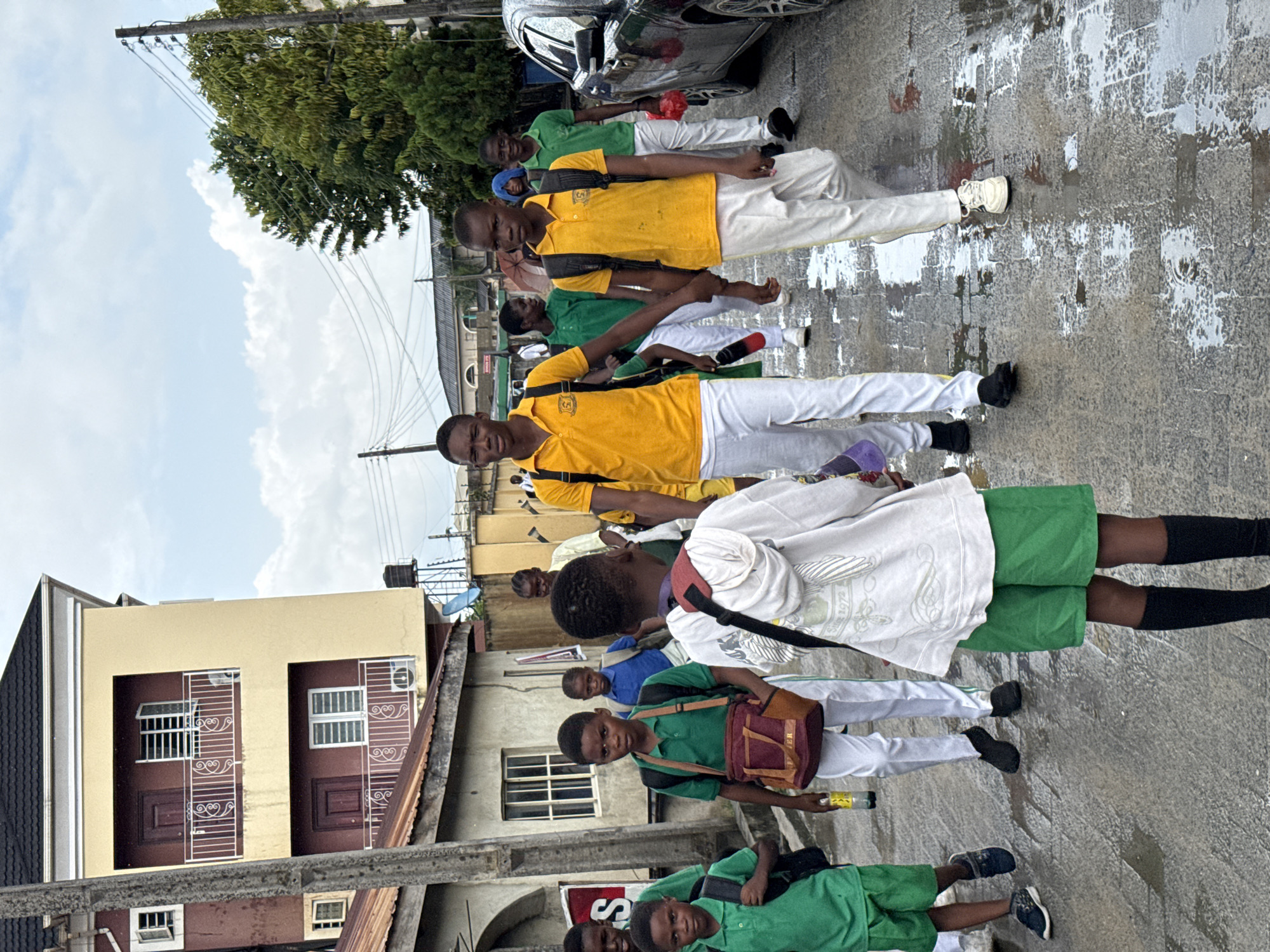 A line of students in school uniforms walks along a wet street in Ado, Nigeria. The students are wearing green and yellow shirts, and white pants. Some students have backpacks. The street is lined with buildings and there are trees visible. A silver car is parked on the right side of the frame. Electrical poles and wires are overhead. The sky is cloudy.