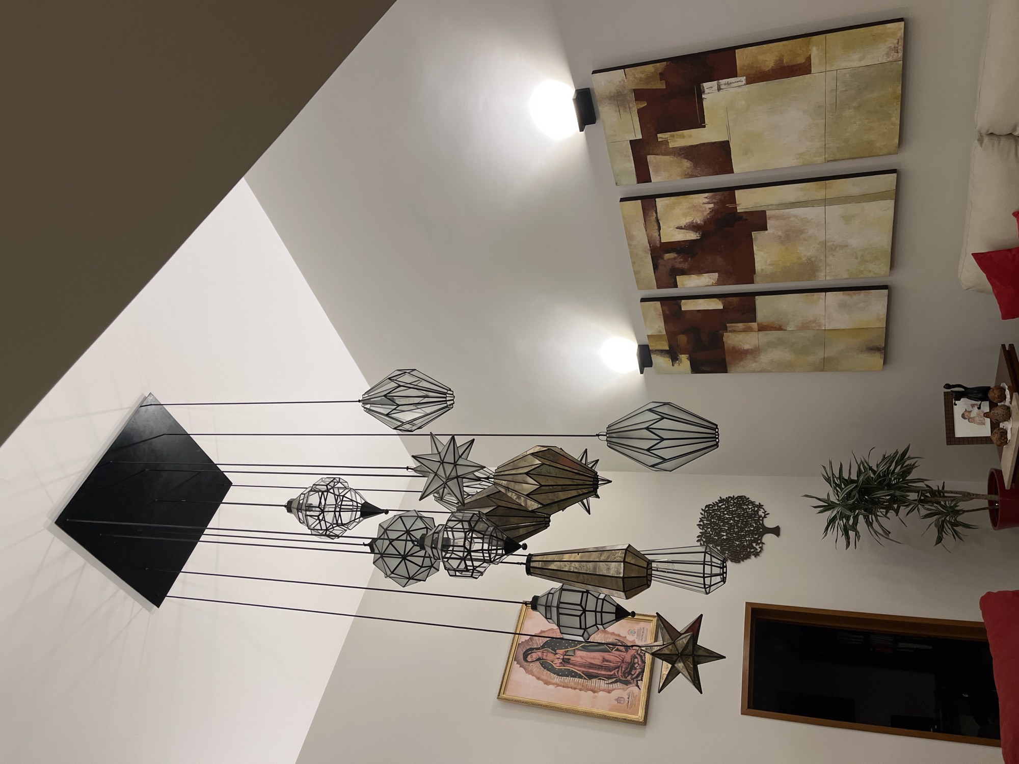 The photograph depicts an interior space in Barrio de Nuevo León, Mexico. A modern chandelier with multiple geometric light fixtures hangs from a square black base attached to the ceiling. The lighting fixture is the most prominent object in the room. The walls are painted white, and the ceiling has an angular design. Three abstract art pieces adorn the wall, and two light fixtures above them. Other objects in the scene include a plant, a framed portrait of the Virgin Mary, and part of a television set. The setting suggests the interior of a residential property.