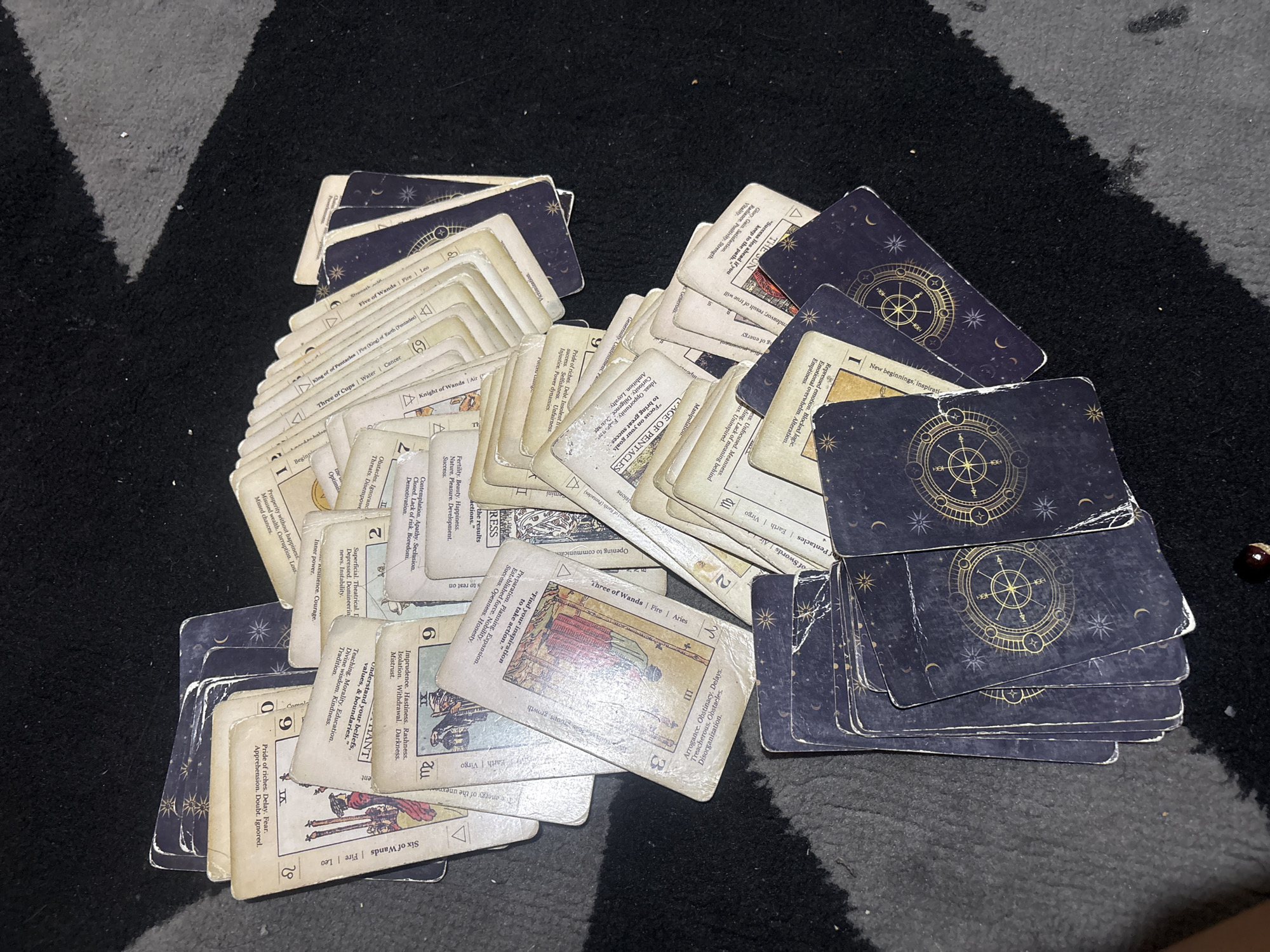 The image displays a collection of numerous tarot cards spread across a dark, textured carpet. The cards are arranged in various overlapping piles and scattered configurations, showing signs of frequent use with visibly worn edges.
Many cards are face down, revealing their dark blue/purple backs, which are adorned with a repeating gold celestial design featuring stars, crescent moons, and a central circular compass-like symbol. Several cards are face up, exposing their fronts, which appear to be from a tarot deck influenced by the Rider-Waite tradition, characterized by aged, off-white card stock and distinct illustrations.
Visible card titles and associated text include "THE SUN" with phrases like "Radiance. Positivity. Strength," "PAGE OF PENTACLES" with "Focus on your goals to bring great success," "Six of Wands," and "Three of Wands." Other decipherable cards include "King of Pentacles," "Three of Cups," "Knight of Wands," "Five of Wands," and cards with numeric designations (e.g., '2', '6', '7', 'I', 'XX'). Each face-up card typically lists its name, elemental and astrological associations (e.g., "Fire | Aries"), and keywords or interpretive phrases for both upright and potentially reversed meanings. A small, dark, bead-like object is visible on the carpet near one of the card stacks. The scene suggests an indoor setting, possibly after a tarot reading or during the handling of the deck.