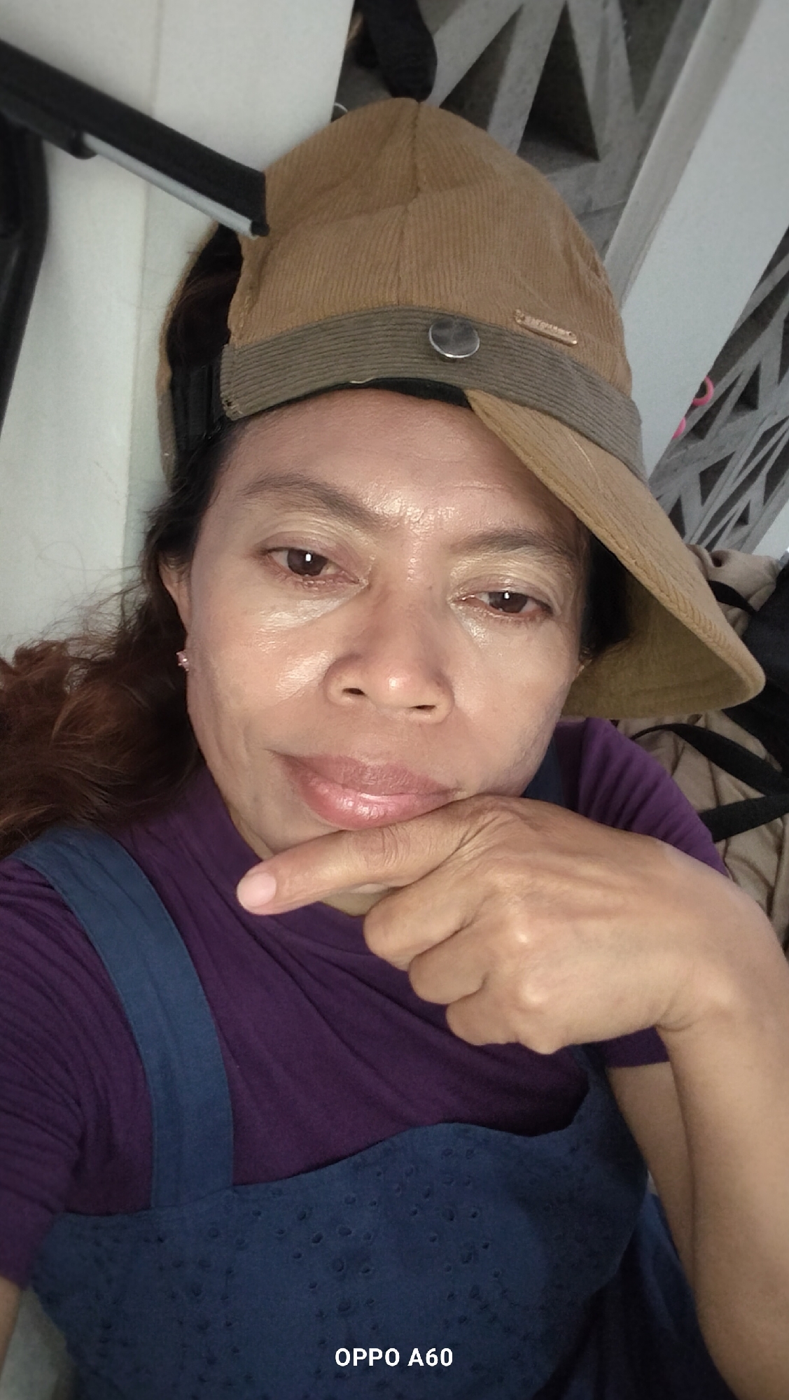 A woman wearing a brown corduroy hat and a purple shirt with blue overalls is looking at the camera. Her right hand is resting on her chin. The photo appears to be taken indoors, possibly in a residential setting in North Kuta, Indonesia. The background is blurred but suggests a domestic environment with what looks like a white wall and some angular structures. The lighting is somewhat harsh, highlighting the texture of her skin and the fabric of her clothing. A watermark at the bottom right corner of the image reads "OPPO A60".