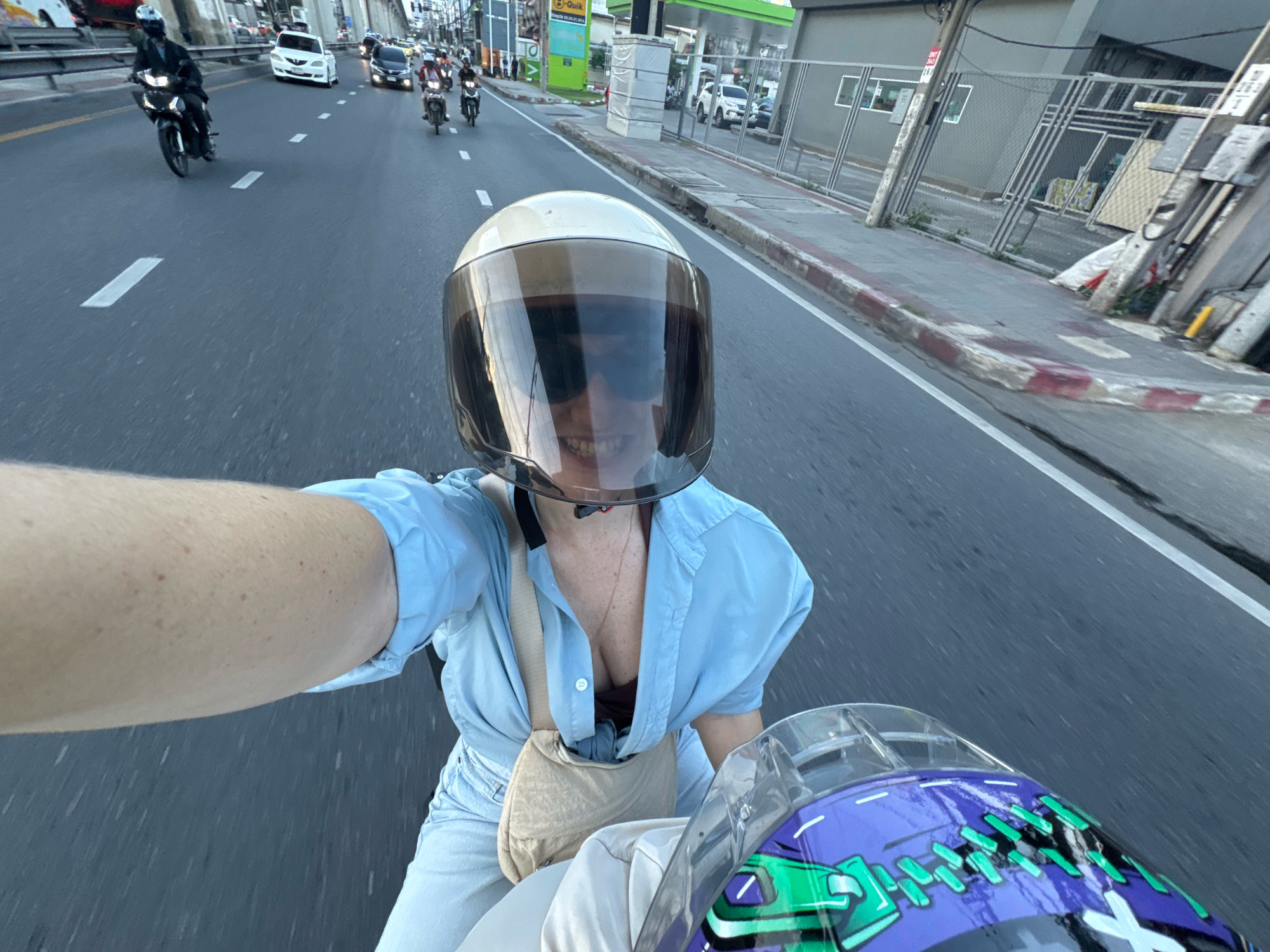 A selfie taken from the perspective of someone riding a scooter in Samrong Nuea Subdistrict Municipality, Thailand. The rider, wearing a blue shirt, light blue pants, and a white helmet with a tinted visor, is smiling. A beige crossbody bag is slung over their shoulder. In the foreground, the front of another scooter, decorated with purple and green graffiti, is visible. The background shows a busy street with other vehicles, including motorcycles and cars. Buildings and a gas station with green signage are visible along the roadside. The road is paved with asphalt and marked with white lane lines. The scene depicts everyday urban traffic in Thailand.