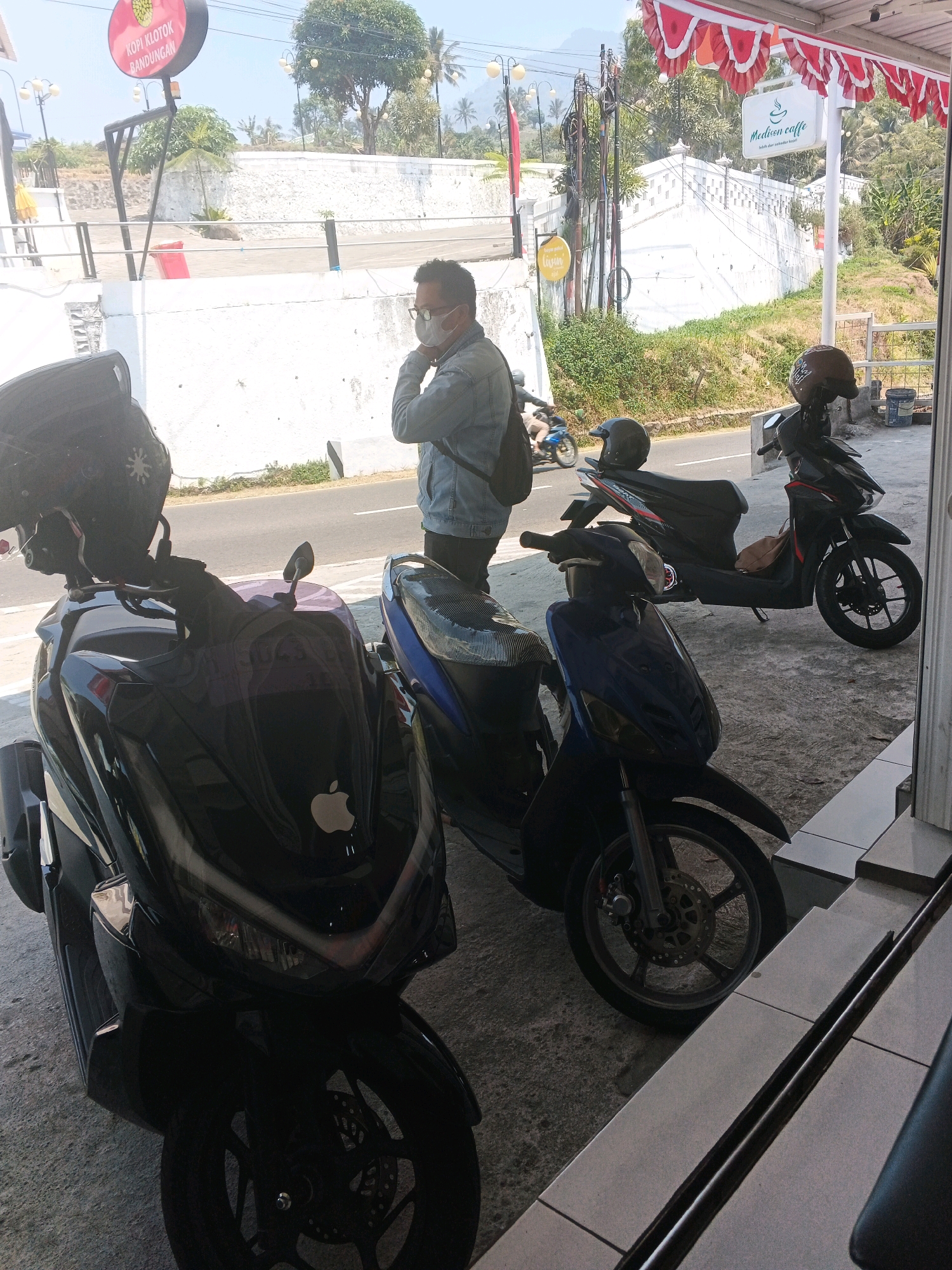 The image shows an outdoor scene in Pakopen, Indonesia, likely near a street. In the foreground, there are two parked scooters, one black and one blue. The black scooter has an Apple logo on its front panel. Behind the scooters, a man wearing glasses and a face mask is standing and looking to his left. He is wearing a denim jacket and carrying a backpack. In the background, a white wall lines a road, and beyond that, there are trees and hills. A red and white banner is strung across the top of the frame. Signs for "Kopi Klotok" and "Madison Caffe" are visible in the background, indicating a commercial area. The overall atmosphere is bright and sunny.