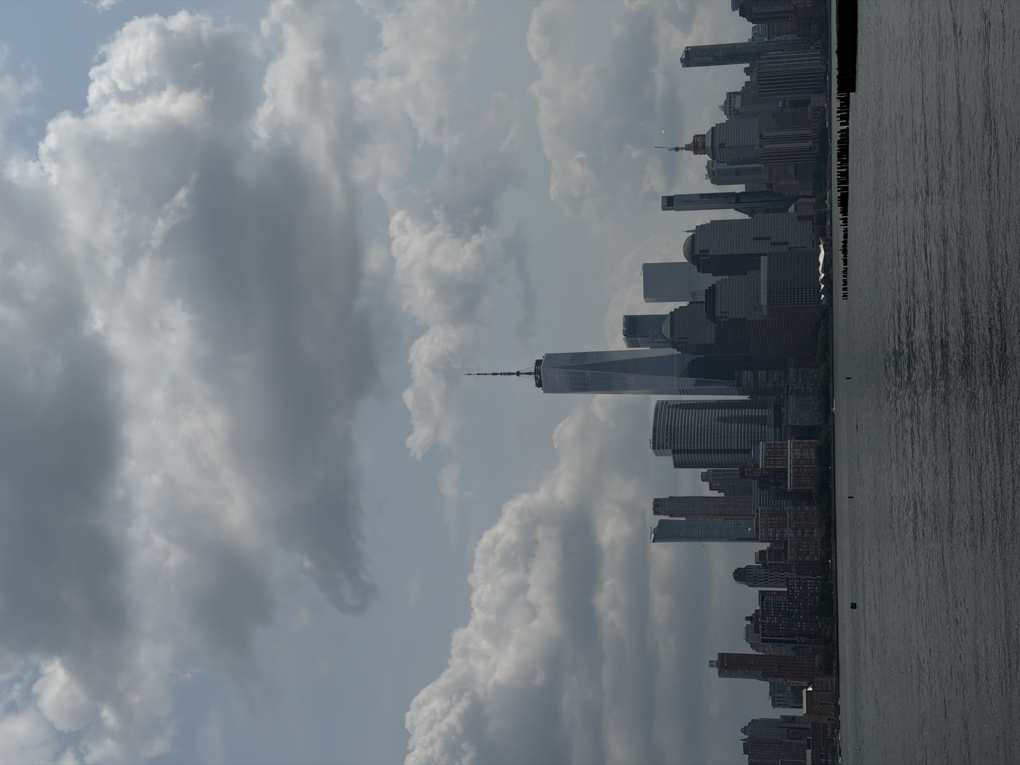 A wide shot captures a New York City skyline with a body of water in the foreground. The skyline is dominated by tall skyscrapers, including the distinctive One World Trade Center. The sky is filled with dramatic, dark clouds, suggesting an overcast or impending storm. The water in the foreground appears choppy and dark. The scene is an outdoor cityscape.