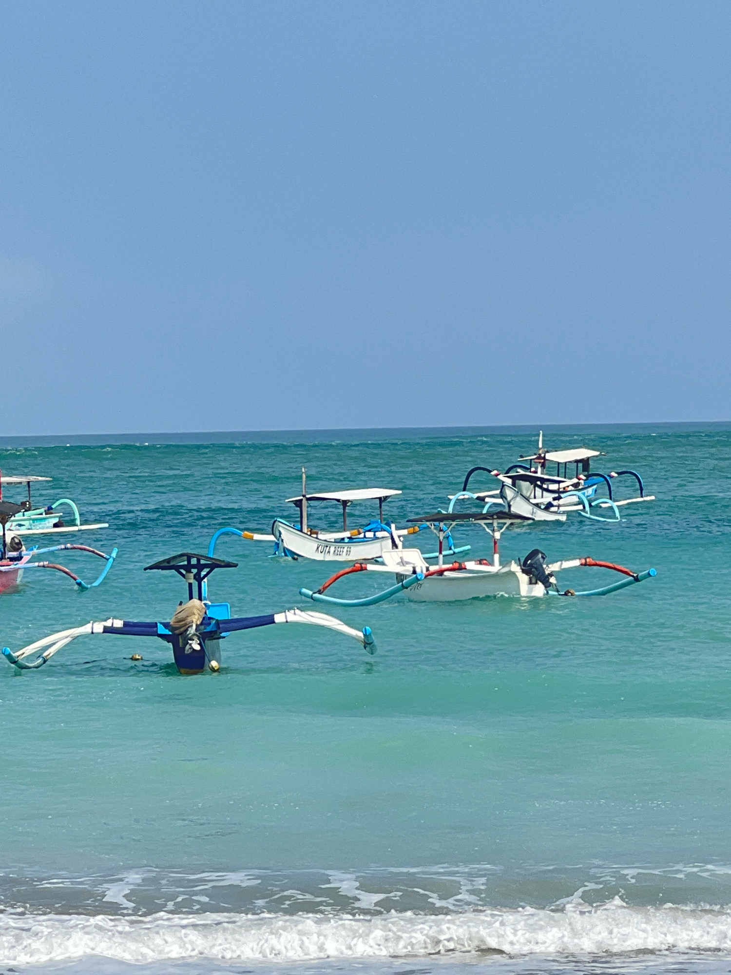 A bright, clear day is captured in this image taken in Kuta, Indonesia. The scene features the turquoise waters of the ocean with several traditional Balinese fishing boats, known as jukungs, dotting the water. These boats are characterized by their outriggers, which extend horizontally from the sides to provide stability. One jukung in the foreground has a person in it, facing away from the camera, with their arms spread wide and resting on the outriggers. The boats are of various sizes and colors, with some having canopies for shade. The sky above is a uniform light blue, and gentle waves are lapping at the shore in the foreground. The overall impression is one of a peaceful coastal scene.