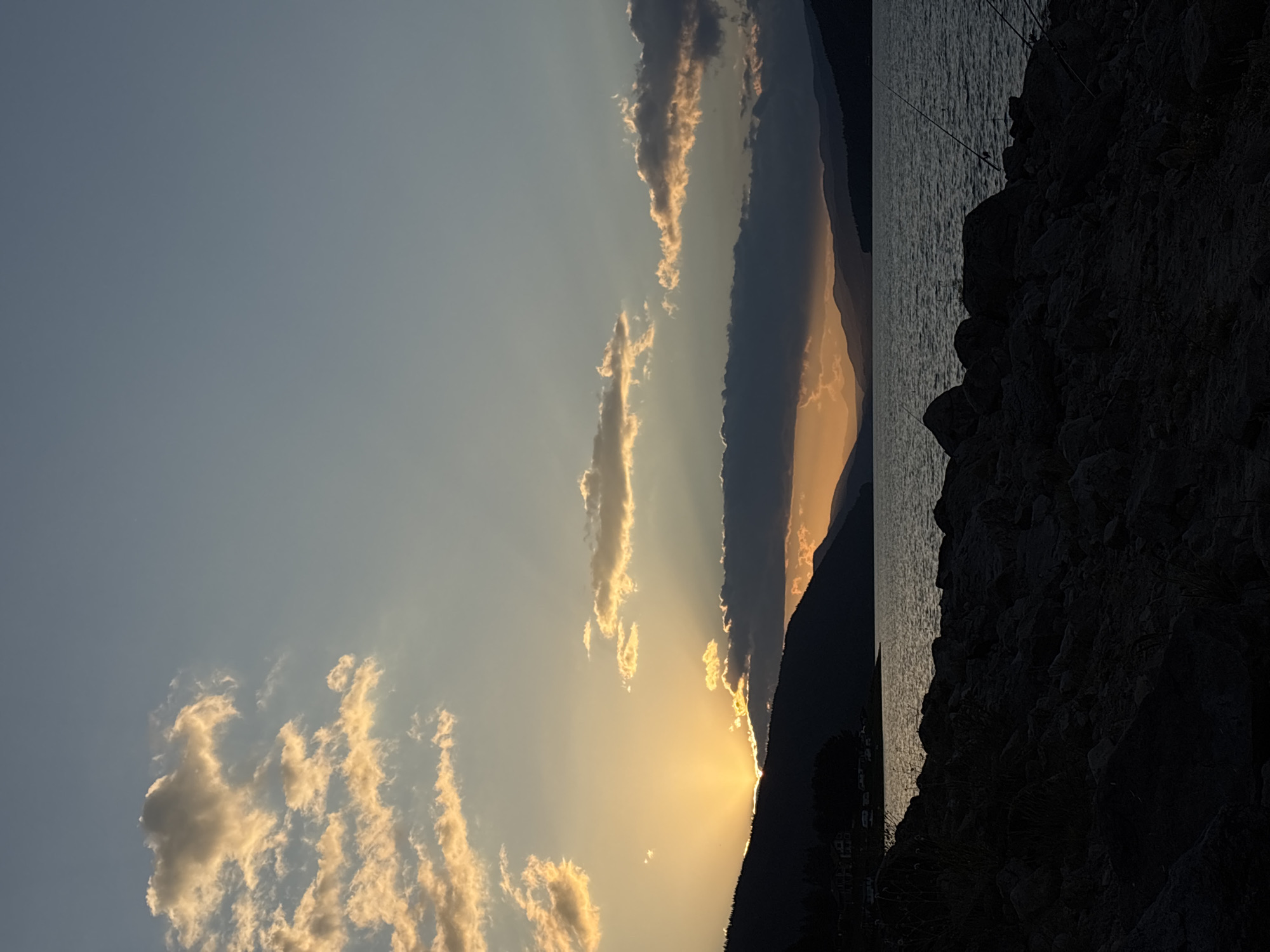 A vertical, low-angle shot captures a sunset over a body of water. The sky is a gradient of blue and orange, with scattered clouds tinged with yellow from the setting sun. The sun itself is partially obscured by a dark cloud formation on the horizon, casting rays of light through gaps in the clouds. In the foreground, a rocky shoreline is silhouetted against the water, with some sparse vegetation growing between the rocks. The water is calm, with gentle ripples reflecting the colors of the sky. Further in the distance, dark mountains line the opposite shore. Several fishing rods are visible on the right side of the frame, planted in the rocks on the shoreline, indicating recreational activity. The overall scene evokes a sense of tranquility and natural beauty.