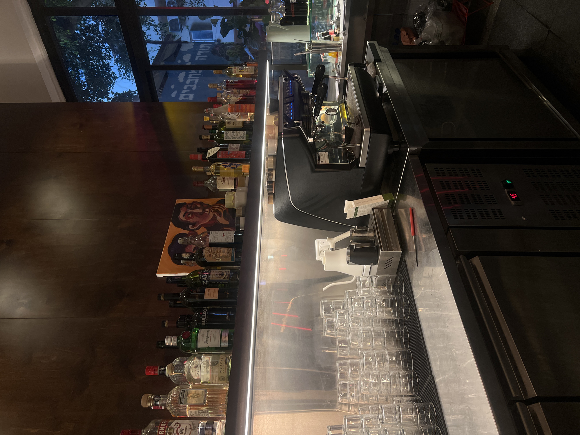 The image shows a bar in Tel Aviv, Israel. A variety of alcohol bottles line the back wall, above an array of stacked glasses. A modern espresso machine sits on the countertop next to the glasses. A refrigerator unit is visible on the right side of the countertop. Through the large window, a partial view of the city is visible. Hebrew text is visible on an outside wall.