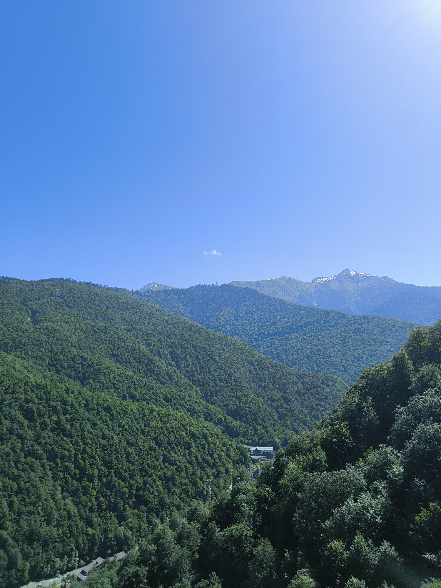 A wide, high-angle outdoor shot shows a lush green mountain landscape under a clear blue sky. The mountains are covered in dense trees, with some snow-capped peaks visible in the distance. A small building complex and a river can be seen nestled in a valley among the trees in the lower-left portion of the frame. The lighting suggests it is daytime, and the overall scene is serene and natural.