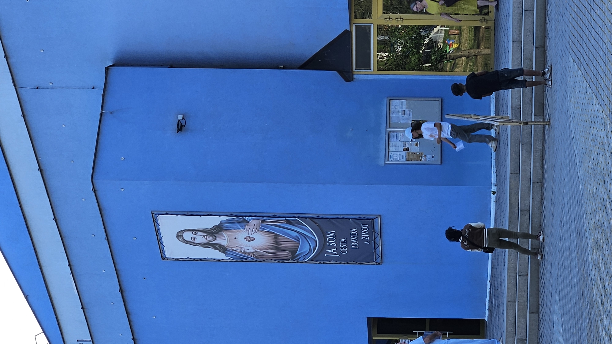 The image shows the exterior of a modern, bright blue building, likely a church or religious community center, in Prešov, Slovakia. Dominating the facade is a large vertical banner featuring an image of Jesus Christ with his hands outstretched, displaying his Sacred Heart. Below the image, the banner displays text in Slovak: "JA SOM CESTA PRAVDA A ŽIVOT," which translates to "I am the way, the truth, and the life."
Several individuals are visible on and around the building's entrance steps. On the steps, two people are standing, one facing away from the camera wearing a brown and cream jacket and dark pants, and another person, partially obscured, in a light-colored outfit. Further to the right, a person in a white t-shirt and grey pants is walking down the steps while holding a piece of paper. Another young person in a black t-shirt and denim shorts stands on the paved area in front of the steps, facing away from the camera.
Adjacent to the entrance, there's a bulletin board with several notices posted. To the right of the bulletin board, a yellow-framed doorway leads into the building, with trees and foliage visible beyond. A woman in a yellow dress is seen walking away from the entrance. The overall environment suggests an outdoor public space during daytime, with a clear blue sky overhead and the building's vibrant blue color reflecting the sunlight.