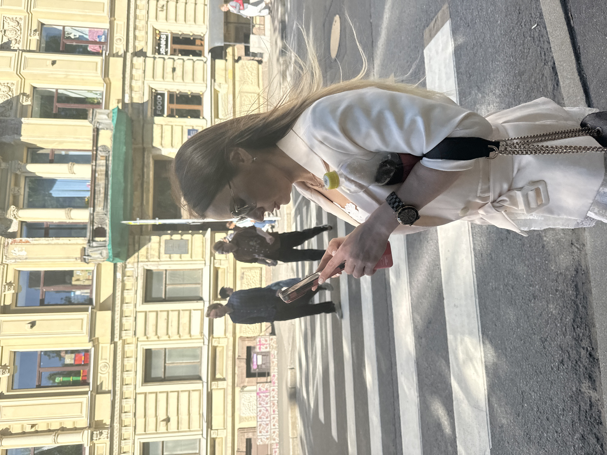 A woman is walking across a pedestrian crossing in Saint Petersburg, Russia, holding a smartphone. She is wearing a white jacket and sunglasses. Her hair is long and brown, and she has a watch on her left wrist. The woman appears to be looking at her phone, which has a red case. In the background, there are buildings with ornate facades and several other people walking. The time of day appears to be midday, with bright sunlight casting shadows. The weather seems to be clear. The scene is bustling with activity, typical of a city street. No visible text is present on the woman or her accessories.