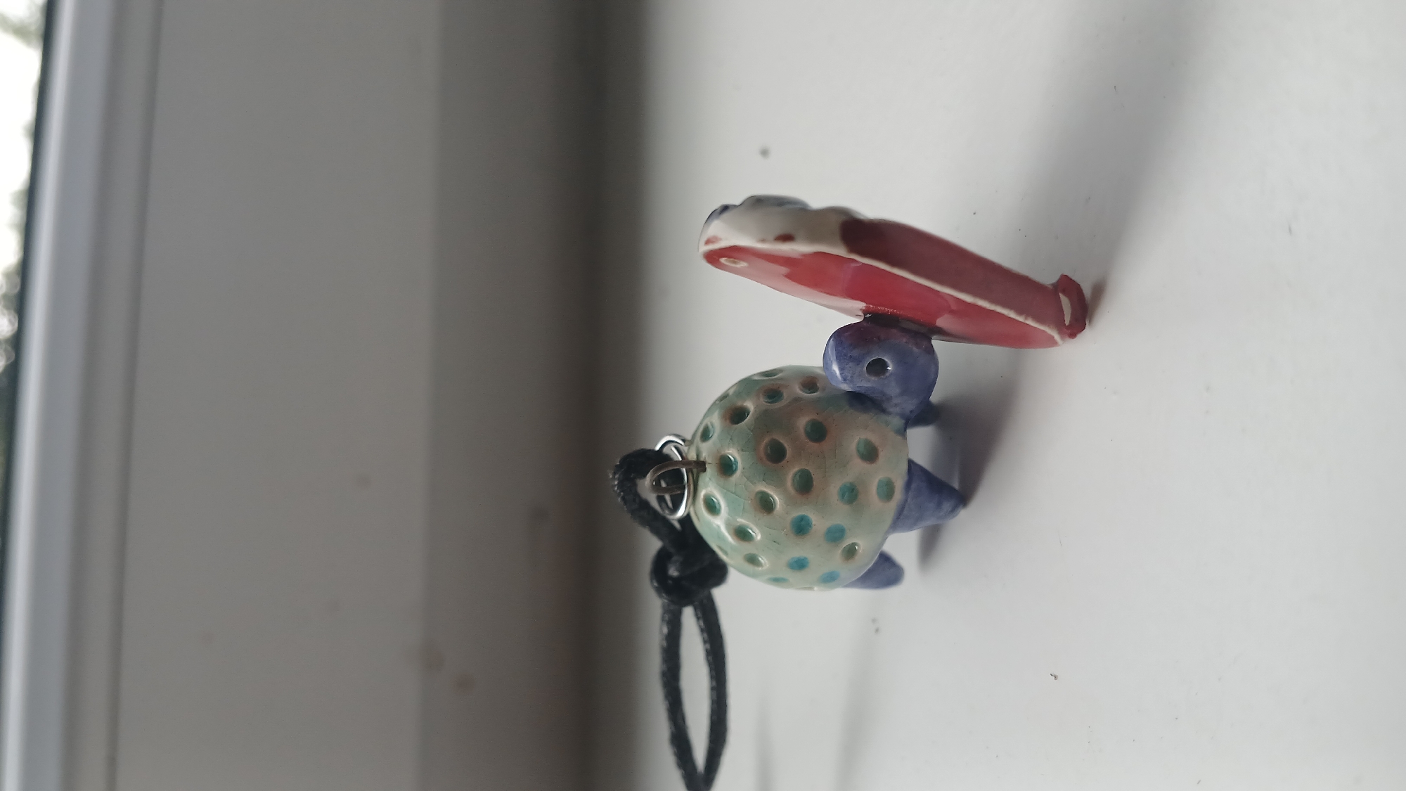 An artistic, ceramic turtle pendant with a light blue-green speckled shell, purple legs, and head sits on a white surface. A red and white round object, possibly a miniature surfboard, is attached to the turtle's front right leg. A black cord is attached to a small metal loop on the turtle's shell. The background is blurred, showing a white ledge and a hint of greenery. The image appears to be taken indoors, possibly on a windowsill. This is not located in Visaginas, Lithuania.