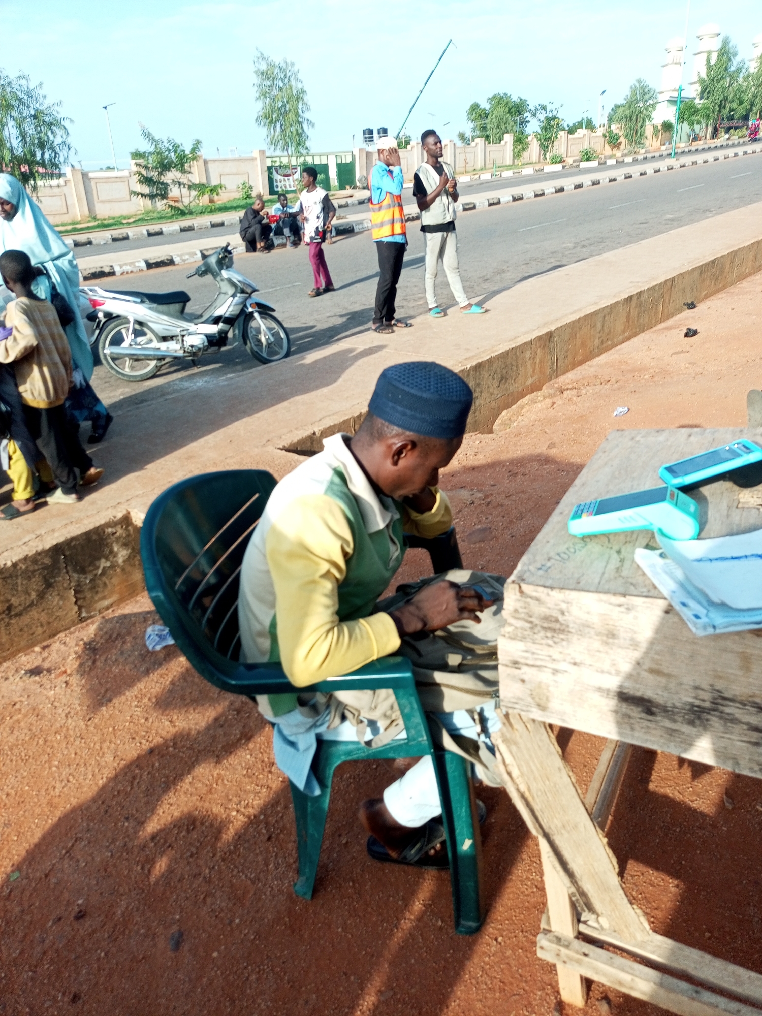 In the city of Gombe, Nigeria, on a sunny day, a man wearing a blue skullcap and a yellow and green shirt is sitting in a green plastic chair on the side of a paved road. He is looking down at a brown bag on his lap, and his right hand is reaching into the bag. In front of him is a rustic wooden table with three teal-colored devices on it, two of which appear to be cell phones. A white cloth is folded on the table. To his left, a silver motorcycle is parked on the side of the road.
Several people are standing and walking along the sidewalk behind the motorcycle. One person is wearing a light blue hijab. Another person, a child, is wearing a tan shirt and yellow pants. Two individuals are dressed in casual clothing, and one is wearing an orange and yellow safety vest over a black shirt and black pants, holding a long thin object. The road is paved and has white lines marking the lanes. The background features a low, light-colored wall, trees, and a white building with domes, likely a mosque. The ground in the foreground is covered in reddish-brown dirt and pebbles. The sky is clear and blue.
