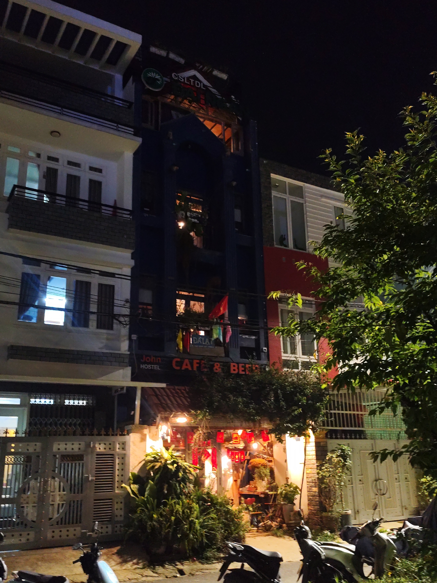 This nighttime image depicts a bustling street scene in Da Lat, Vietnam, characterized by multiple multi-story buildings and vibrant artificial lighting. The central focus is a tall, dark blue building housing "John HOSTEL" and a "CAFE & BEER" establishment, as indicated by illuminated signs above its ground floor. A smaller sign reading "DALAT" is also visible near a window on this building. Higher up, a circular green logo with a plant design and the letters "CSLTDL" is affixed to its facade.
To the left, a white building with several balconies and brightly lit windows stands in contrast. To the right, a building featuring a red base and white upper stories is partially obscured by a dense green tree. The ground level of the cafe/hostel is brightly illuminated with warm lights and decorated with traditional red lanterns and various potted plants, creating an inviting ambiance. Several motorbikes are parked on the street in the immediate foreground. The night sky is dark and clear, with artificial lights casting a glow on the urban environment.