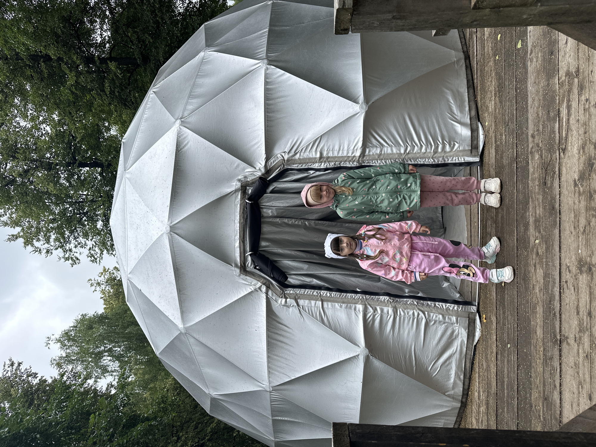 Two young girls stand in front of a geodesic dome structure. The girl on the left wears a pink tracksuit with a Mickey Mouse design and a pink headband. The girl on the right wears a green hooded jacket with a floral pattern and pink leggings. They are standing on a wooden platform in front of the entrance to the dome, which is covered by a gray curtain. The geodesic dome appears to be made of a silver, reflective material and is surrounded by trees. The sky is overcast. The scene is outdoors, and the ground is covered with wooden planks and scattered leaves. This image was taken at the Udmurt Republic Museum of Fine Arts in Izhevsk, Russia, which has a geodesic dome exhibit.