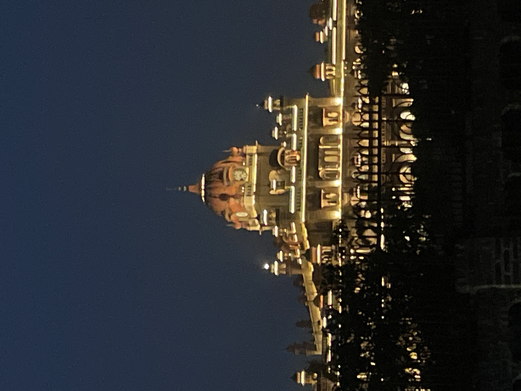 The image shows a large, ornately decorated building illuminated at dusk, situated in Amritsar, India. The building, likely a palace or significant historical structure, features a prominent dome and multiple turrets, all highlighted with warm, yellow lighting. The architecture appears to be of a traditional Indian style, with intricate detailing visible on the facade, including arched windows and decorative elements. In the foreground, the silhouette of trees and a decorative fence obscures the lower portion of the building, adding a sense of depth and framing the structure. The sky is a deep, clear blue, characteristic of twilight, with no discernible celestial bodies like the moon or stars. The scene is static, with no people or animals visible, focusing solely on the architectural grandeur of the illuminated building against the darkening sky. The overall impression is one of nocturnal beauty and historical significance.