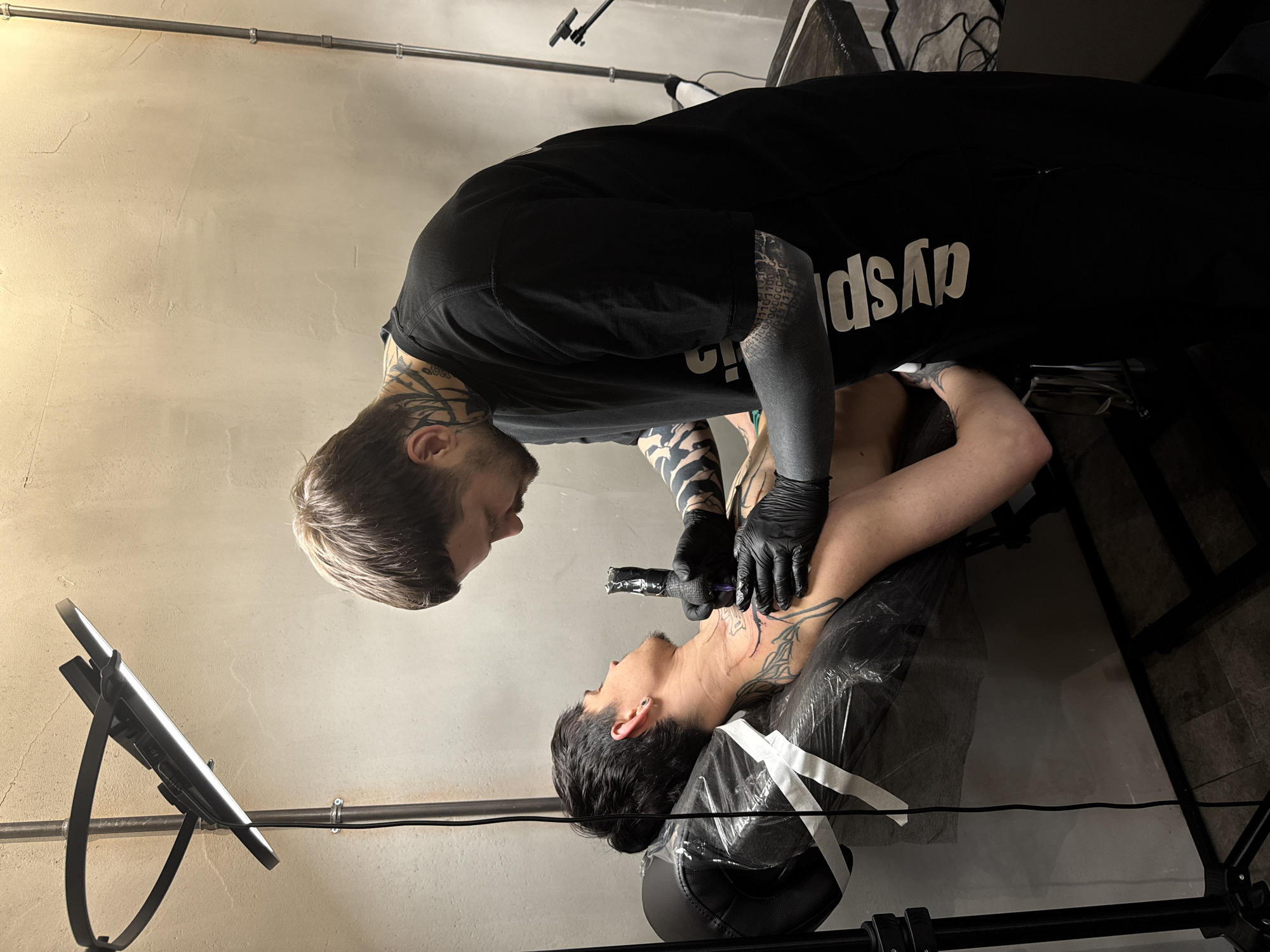 This media file captures an indoor scene within what appears to be a tattoo studio in Moscow, Russia. Two men are featured: a tattoo artist and his client.
The tattoo artist, standing over the client, has a dark beard, light brown hair, and extensive blackwork and tribal-style tattoos covering his arms and neck. He wears a black short-sleeved t-shirt, black protective gloves, and is intently focused on his work. On his t-shirt, the white, reversed text "dysp" is visible. He holds a tattoo machine, carefully applying ink to the client's neck.
The client is lying on his back on a black tattoo bed or chair, his head slightly elevated and tilted, exposing his neck and upper chest. He has short dark hair, a beard, and existing tattoos on his neck and arm. A clear plastic sheet covers his torso and the bed, a standard hygienic practice during tattooing. His expression is neutral, suggesting composure during the session.
The setting features a light-colored, textured wall and a metal pipe structure running horizontally along the upper part of the wall, hinting at an industrial or functional aesthetic. To the left, a tablet or screen on a stand is visible, possibly displaying reference designs or playing entertainment. The lighting is even, indicating an artificially lit indoor environment. The scene depicts a professional and focused tattooing session.
