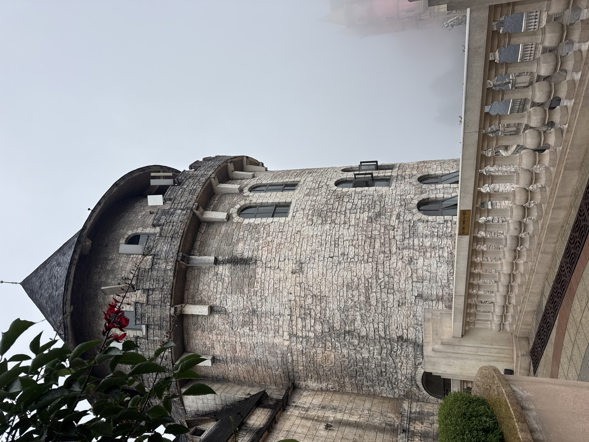 The image depicts a section of the French Village at Sun World Ba Na Hills in Đà Nẵng, Vietnam. A stone tower with a conical roof is the central subject, featuring arched windows and decorative stone ledges. The tower is textured with a pattern resembling stacked stone blocks. To the left, foliage obscures the lower portion of the tower. To the right of the tower, a section of a building with a light-colored balustrade is visible. The upper portion of the image is filled with a gray, overcast sky. The photo is oriented in a manner where the tower appears to be leaning.