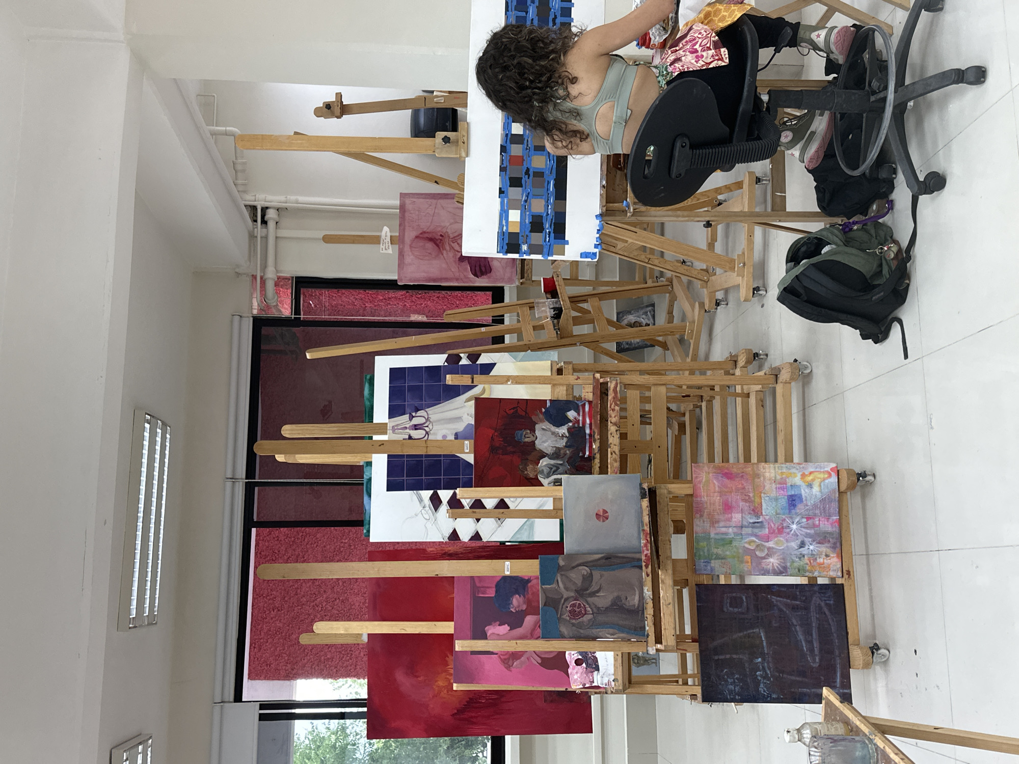 A person with curly hair, wearing a light green top and patterned shorts, sits in a black chair in an art studio in San Andrés Cholula, Mexico. They are facing and working on a painting with a grid of colored squares. Numerous wooden easels support various paintings. Some paintings appear abstract, while others depict figures. A green backpack sits on the floor beside the chair. The room has white walls and what appear to be large windows with dark red blinds. The floor is white tile.