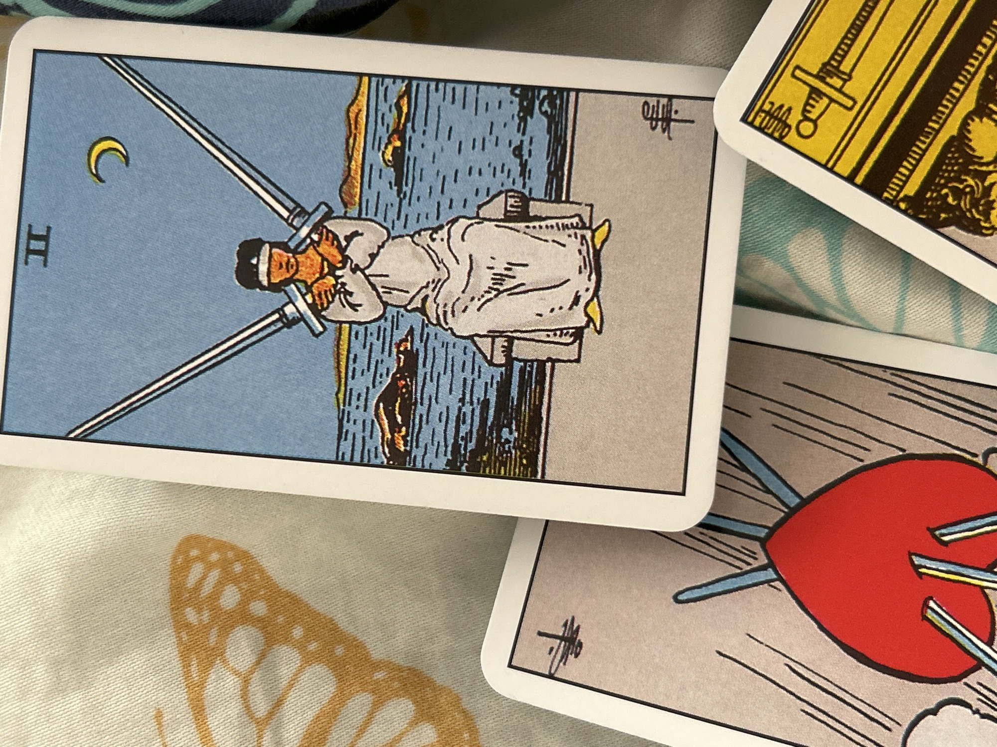 The image shows parts of four tarot cards scattered on a patterned fabric surface. The card most visible is the Two of Swords. It depicts a person sitting on a stone bench, blindfolded, and holding two swords crossed in front of them. Behind the person is a body of water, with a crescent moon visible in the sky. To the right of this card, a section of the Emperor card is visible. Below, part of the Three of Swords card, showing a red heart pierced by three swords, is seen. An unidentifiable card fragment sits below the Two of Swords. The location and city are Unknown.