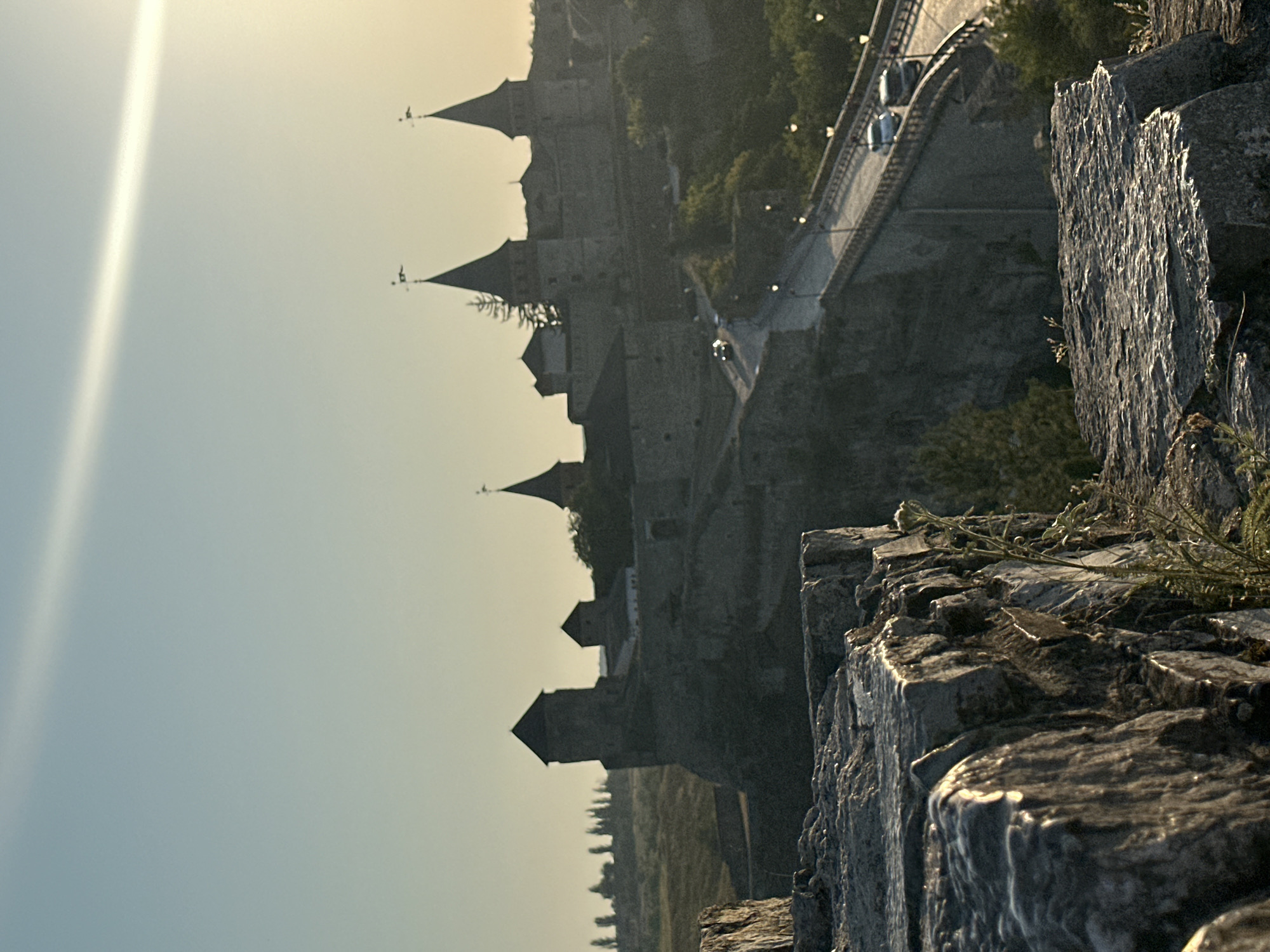 The image captures a panoramic view of the Kamianets-Podilskyi Castle and its surroundings in Ukraine, bathed in the warm glow of late afternoon or early morning sunlight. In the foreground, rough, weathered rocks dominate the lower left and right portions of the frame, their textured surfaces catching the light. Beyond these rocks, the imposing stone structure of the Kamianets-Podilskyi Castle complex unfolds. Several towers with conical roofs, characteristic of the castle's architecture, rise against the pale, clear sky. A bridge, likely the main access route to the castle, stretches across a gorge, with several vehicles, including cars, visible on its surface, suggesting ongoing traffic. Streetlights line the bridge, hinting at its use into the evening. The castle and bridge are nestled within a landscape of rolling hills and vegetation, with trees dotting the background and the faint silhouette of more distant trees against the horizon. The overall scene conveys a sense of historical grandeur and the enduring presence of the fortress amidst a natural setting.