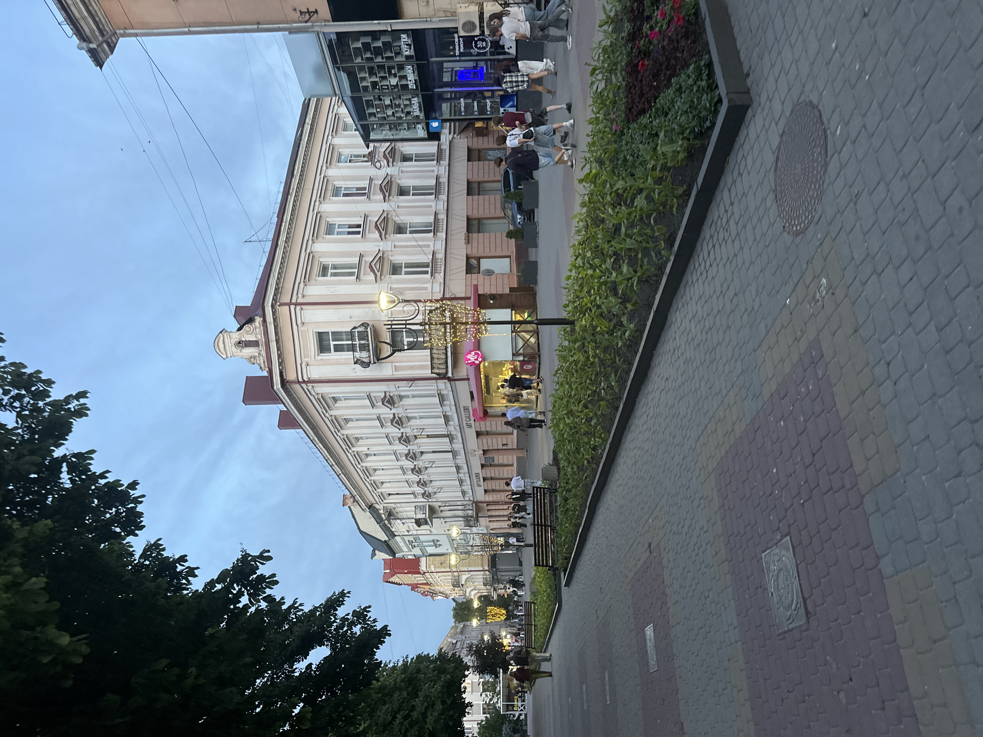 This image depicts a pedestrian street in the city of Ternopil, Ukraine, during the early evening or dusk. The scene is illuminated by soft natural light from the sky and artificial light from streetlights and shop windows.
The foreground features a wide, paved pedestrian area composed of multi-colored rectangular pavers in shades of grey, purple, and muted yellow. Several square and one round metal utility covers are embedded in the paving. Running parallel to the main path are long, narrow flower beds containing low-lying green shrubs and patches of red and purple flowering plants, bordered by dark, low retaining walls. Multiple black metal benches with wooden slats are positioned along these beds.
On the left side of the street, dense, dark green leafy trees extend into the upper left corner, partially obscuring the sky and distant buildings. Further down this side, more multi-story buildings with varied facades are visible, along with additional streetlights decorated with string lights.
The right side of the image is dominated by a row of multi-story historical buildings. The prominent building in the mid-right is light peach-colored with classical architectural details, including ornate window frames, small balconies with iron railings, cornices, and a red-tiled roof with an elaborate gable. The ground floors of these buildings house various commercial establishments, featuring large display windows. Visible Ukrainian signage includes "ART GALLERY" on multiple storefronts, "ШУБИ," "ПАЛЬТА," "ДУБЛЯНКИ," "КУРТКИ" (Furs, Coats, Sheepskin Coats, Jackets) on a clothing store, and "ПРОДАЖ," "ОБМІН," "КРЕДИТ" (Sale, Exchange, Credit) along with an Apple logo and "ТЕРМІНОВИЙ РЕМОНТ" (Urgent Repair) on a glass-fronted electronics/repair shop. A dark-colored sedan or SUV is parked on the street next to these buildings.
Numerous individuals are present on the street, primarily walking. A group of approximately seven to eight younger people, dressed casually in shorts, t-shirts, and jeans, are walking towards the camera in the mid-right. Other individuals and small groups are seen walking in both directions further down the street or standing near shops and benches. The overall atmosphere appears calm and uncrowded.