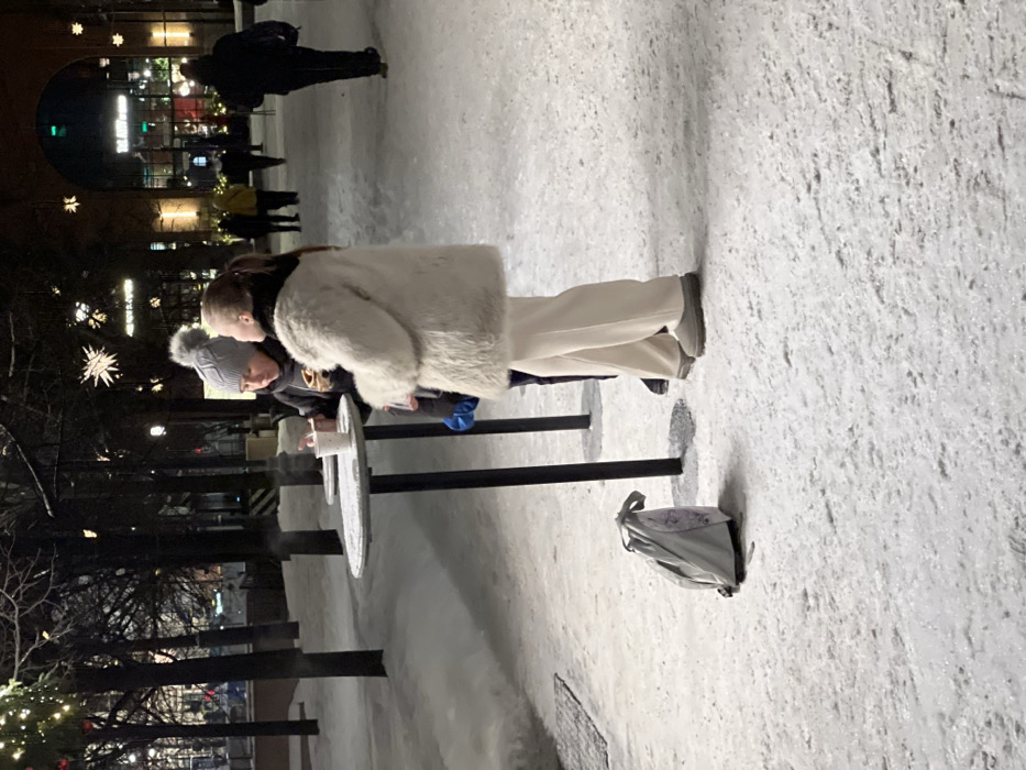 Two people stand near a round table outdoors, with a gray bag resting on the snowy ground beside them. One person wears a white coat and white pants. In the background, several people are walking. A building with lights and decorations stands farther back, and trees are adorned with star-shaped lights. The setting appears to be a winter evening in a public space.