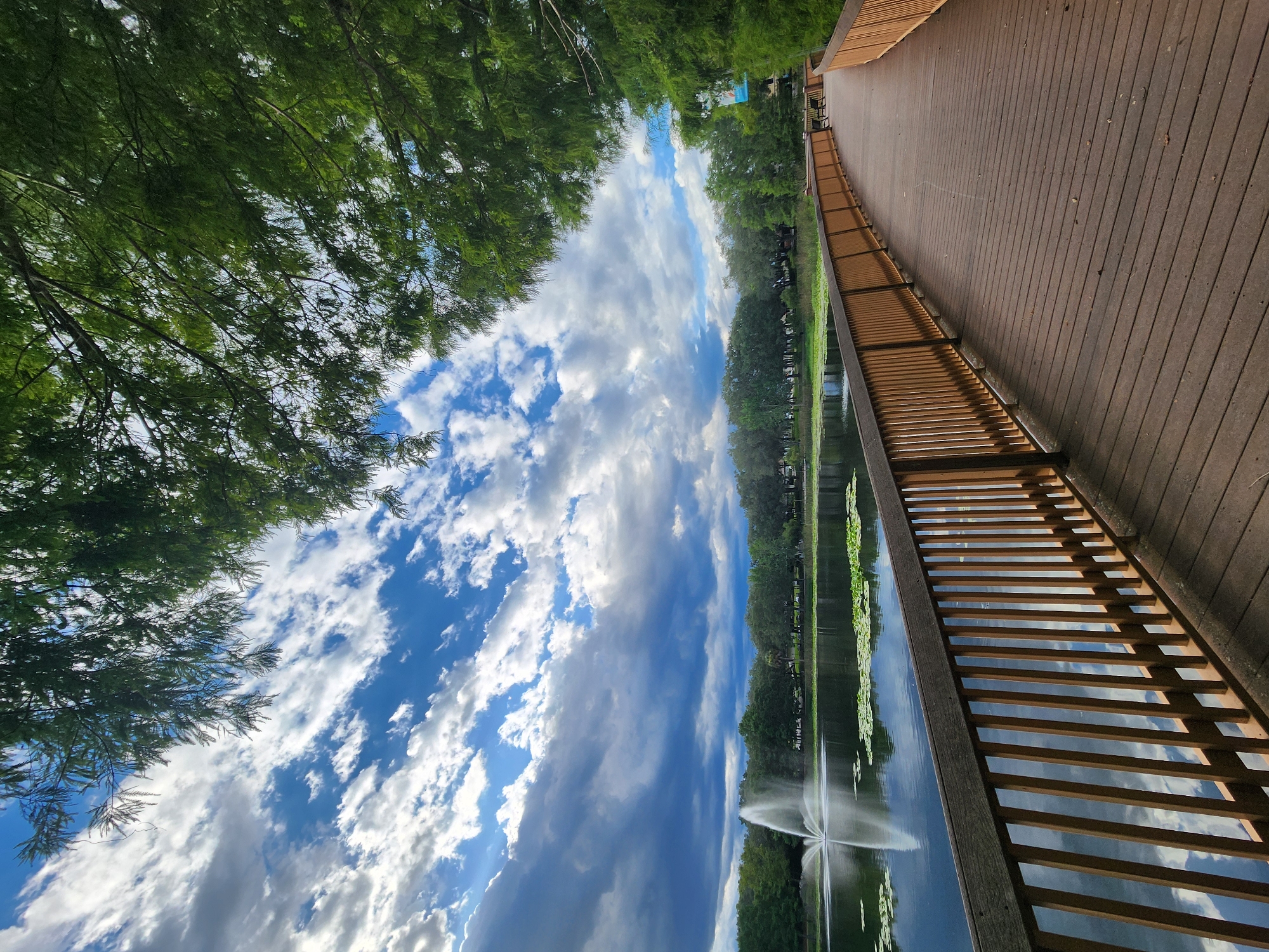 A wooden boardwalk curves along the edge of a lake, its surface reflecting the dramatic sky above. Green trees line the far bank of the lake, with a fountain spraying water in the distance. The sky is a mix of bright blue and heavy, dark clouds, creating a dynamic and somewhat moody atmosphere. The boardwalk is made of dark wood planks and has a light brown wooden railing. The scene appears to be in a park or recreational area in Maitland, Florida.