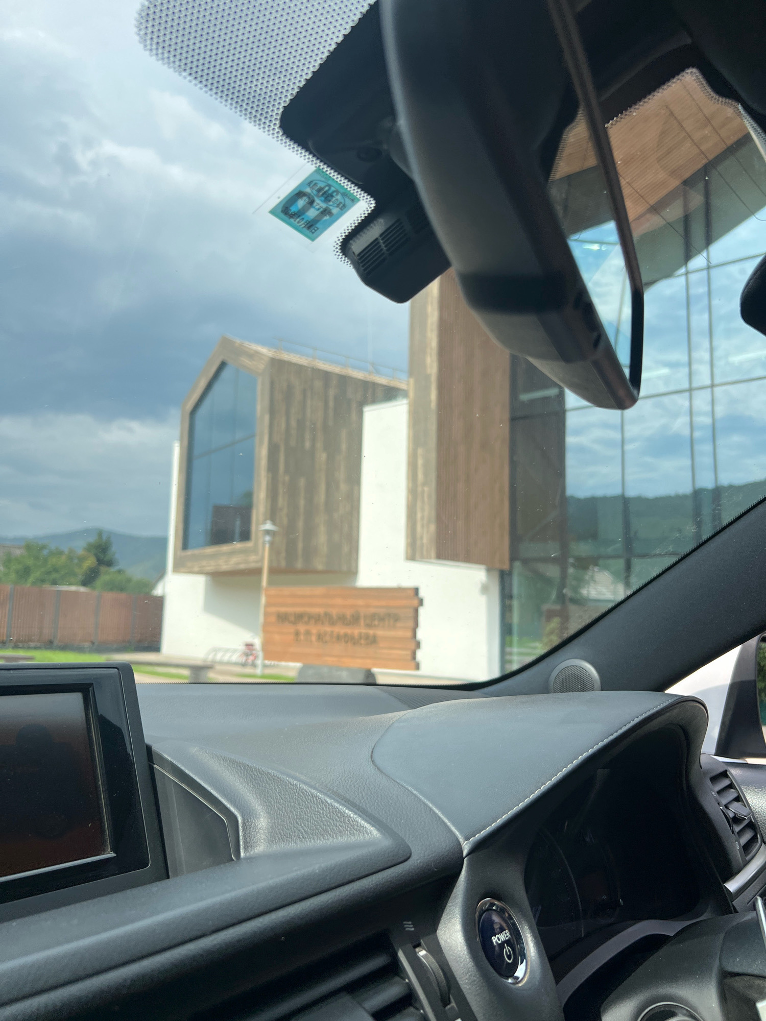 The image is taken from inside a car, looking through the windshield at a modern building with wooden accents. A sign on the building reads "НАЦИОНАЛЬНЫЙ ЦЕНТР В.П. АСТАФЬЕВА" which translates to "NATIONAL CENTER V.P. ASTAFIEV". This indicates the location is the V.P. Astafiev National Center in Ovsyanka, Russia. The sky is overcast with dark clouds. The car's interior is visible in the foreground, including the dashboard, a touchscreen display, and the steering wheel with a power button. A small sticker is visible on the windshield.