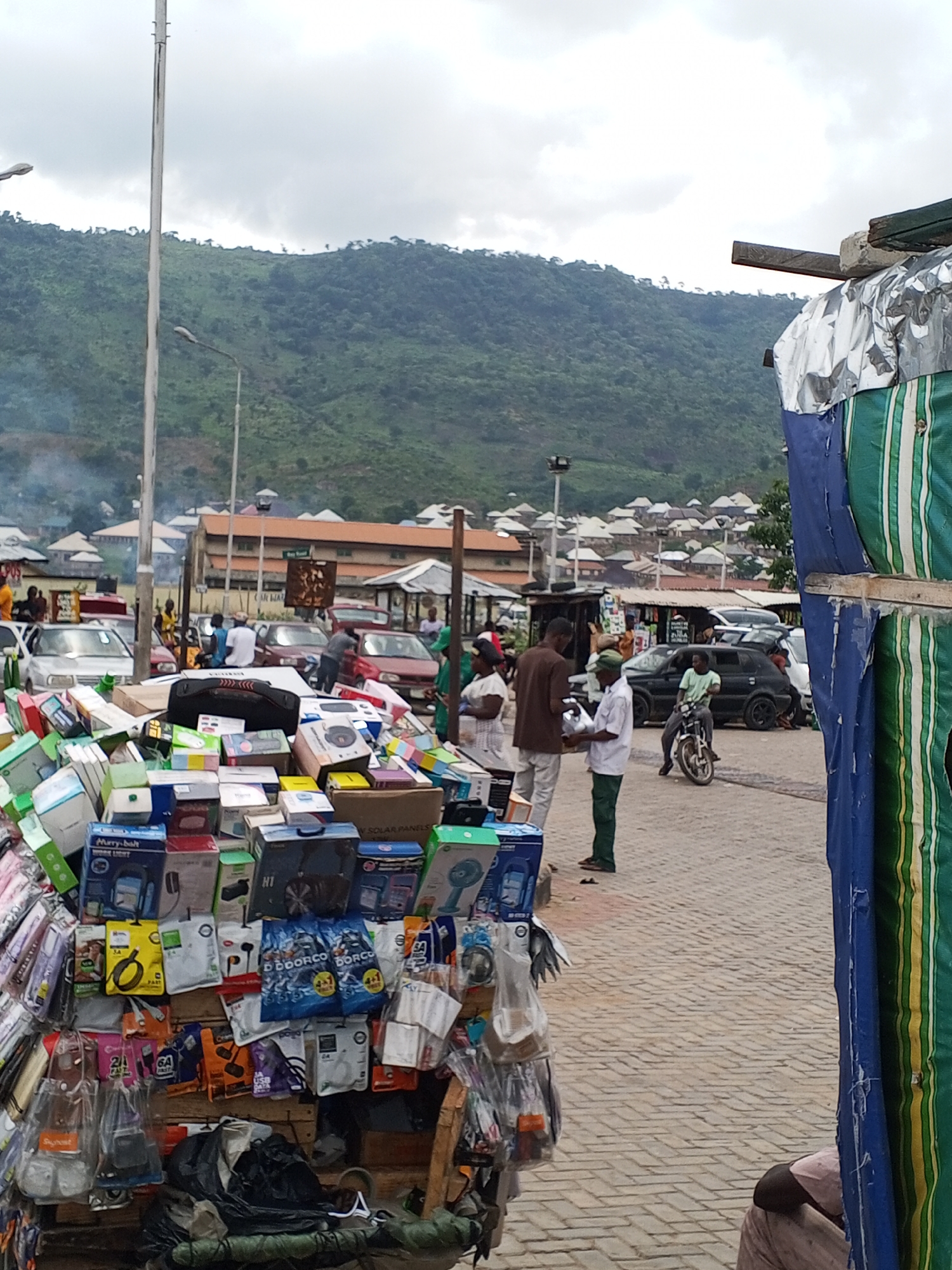 The scene depicts a bustling marketplace in Lokoja, Nigeria. In the foreground, there is a pushcart overflowing with various electronics, including packaged headphones, phone chargers, and small appliances. Behind the pushcart, several people are standing and interacting. Cars are parked along the roadside, and a man is sitting on a motorcycle. In the background are buildings and rooftops nestled against the backdrop of a green, hilly landscape. The sky is overcast, suggesting a cloudy day.