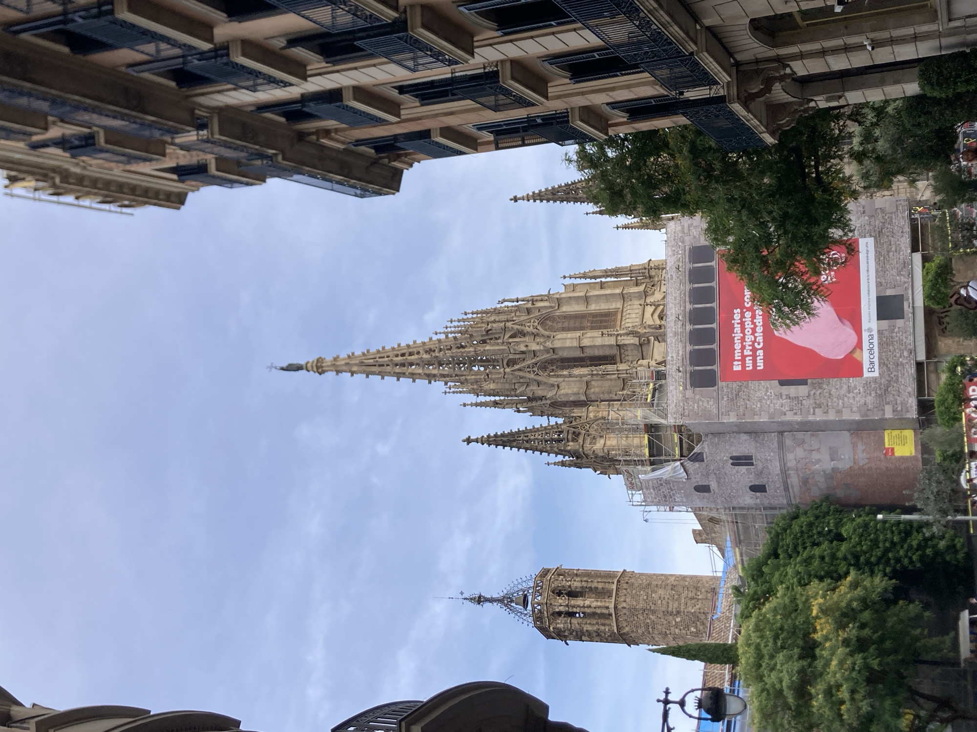 The image depicts a view of the Barcelona Cathedral, also known as the Cathedral of the Holy Cross and Saint Eulalia, located in the Gothic Quarter of Barcelona, Spain. The cathedral's spires and a bell tower are visible against a cloudy sky. The facade of the cathedral is partially covered by scaffolding and a large red advertisement banner featuring a pink ice cream and text that appears to be in Catalan. Adjacent to the cathedral, to the right, is a multi-story building with balconies. To the left of the cathedral, a portion of another building is visible, and some greenery, including trees, is present in the foreground. The scene suggests that the cathedral might be undergoing renovation or restoration. The overall lighting suggests it is daytime, possibly overcast.