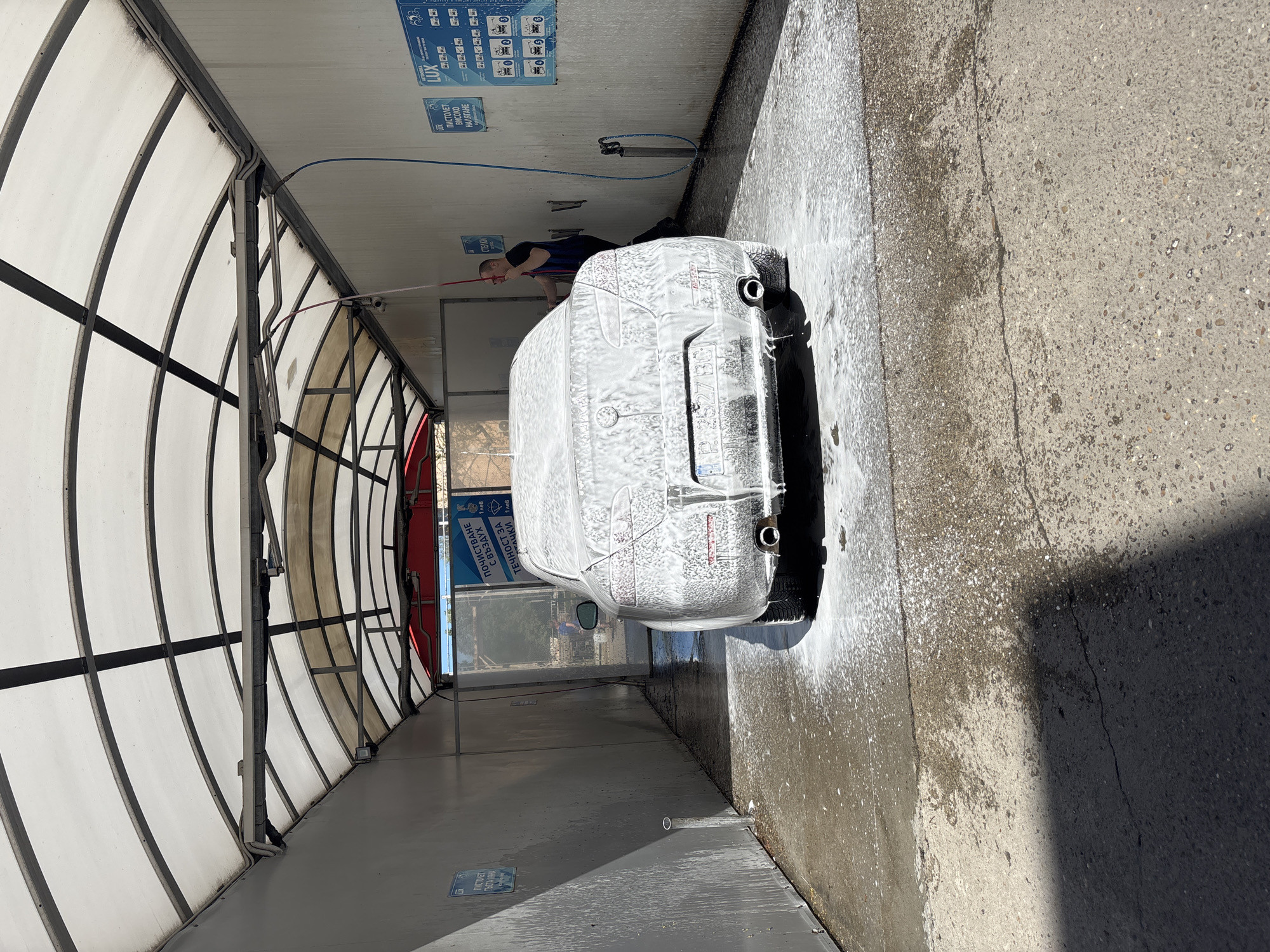 The image shows a white car covered in soap at a self-service car wash station. A man, dressed in dark clothing, is seen using a pressure washer to clean the vehicle. The car wash area is enclosed by a structure with a white, translucent roof and metal framing. On the wall of the car wash, there are blue signs with instructions for using the different cleaning options. The ground is wet from the cleaning process, and there is foam runoff. The sign text suggests this location is in Bulgaria. Specifically, the sign lists LUX and "Пиистолет Високо Налягане" which translates to "High Pressure Pistol" in Bulgarian, plus "Течности за Джанти и Стелки" which means "Wheel and Floor Mat Cleaning". Based on the sign, the location is likely Ruse, Bulgaria.