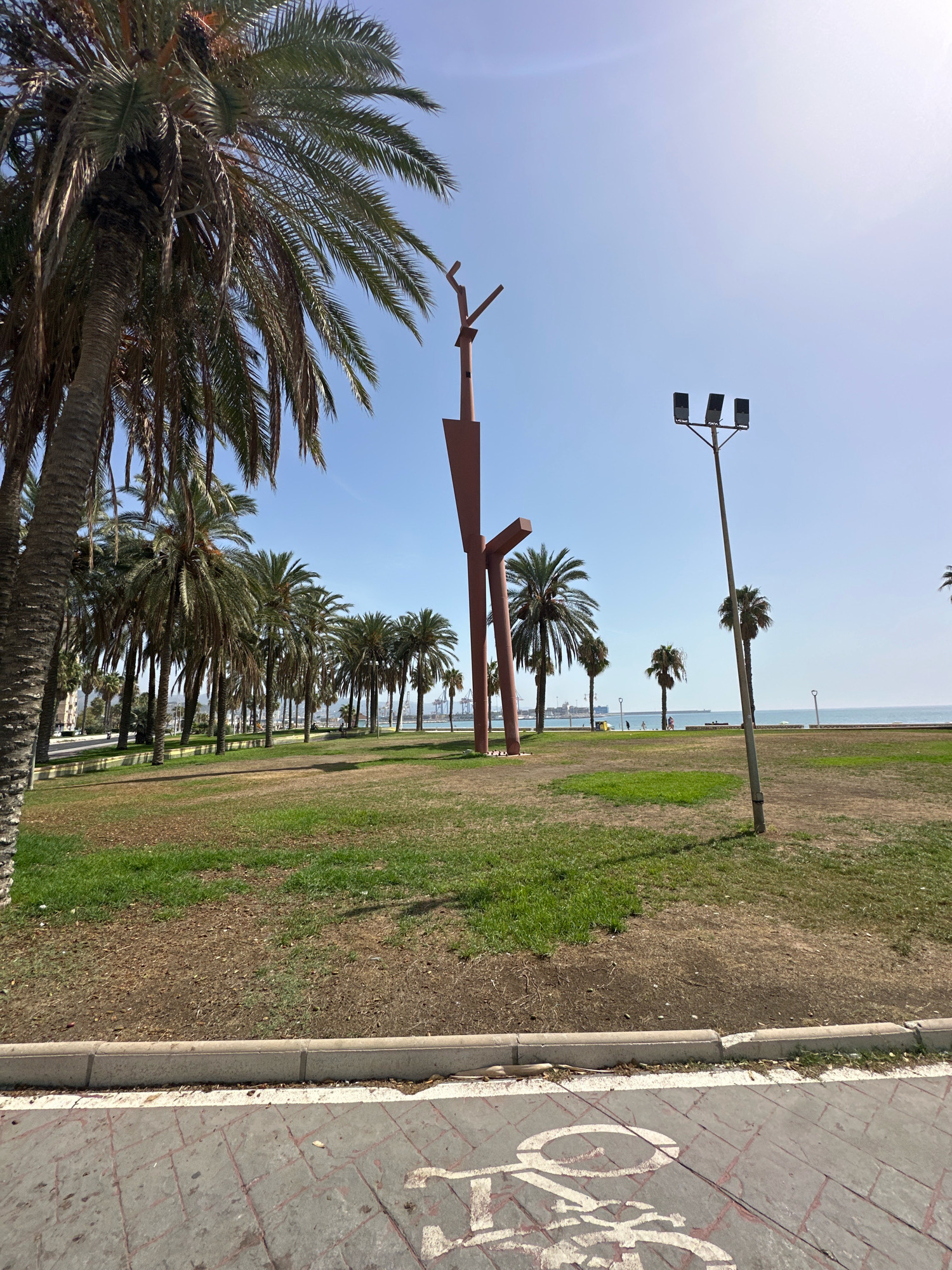 A tall, reddish-brown abstract sculpture stands in a park with palm trees and a view of the sea in Málaga, Spain. The sculpture is made of metal and has a vertical element with two horizontal arms extending from it. In the foreground, a paved path with a bicycle lane marking is visible. Beyond the sculpture and trees, the sea stretches to the horizon, with a few boats in the distance. The sky is clear and blue. The park is green with patches of dry grass. A tall lamppost with three lights stands to the right of the sculpture.