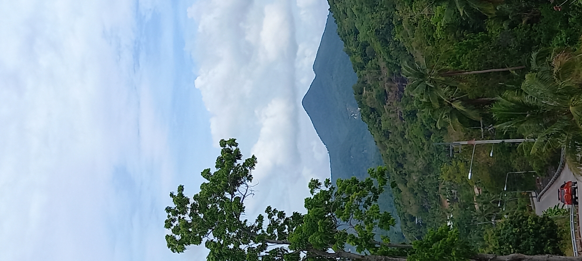 The image captures a landscape in Baan Tai, Thailand. A prominent, slightly off-center mountain dominates the backdrop, partially obscured by fluffy white clouds. The mountainside is covered in dense, verdant tropical vegetation. In the foreground, the branches of a green tree reach into the frame. A curved road with utility poles and streetlights runs along the base of the mountain on the right side of the image, with a glimpse of a red vehicle partially visible. The sky above is a pale blue, filled with scattered clouds.
